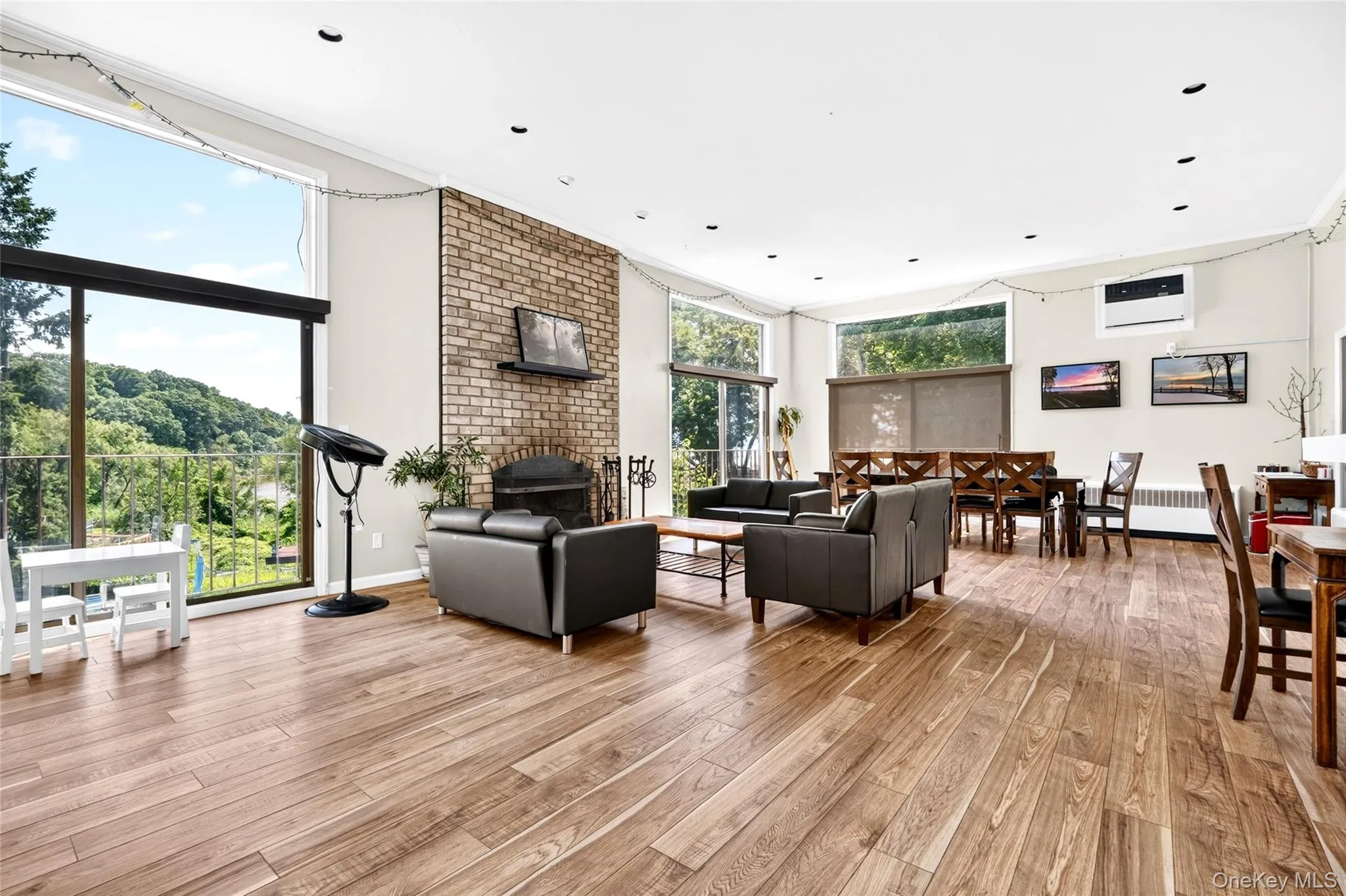 Spacious open-concept room featuring wide-plank wood-finish flooring, a brick-front fireplace, and large windows with outdoor access Spacious open-concept room featuring wide-plank wood-finish flooring, a brick-front fireplace, and large windows with outdoor access
