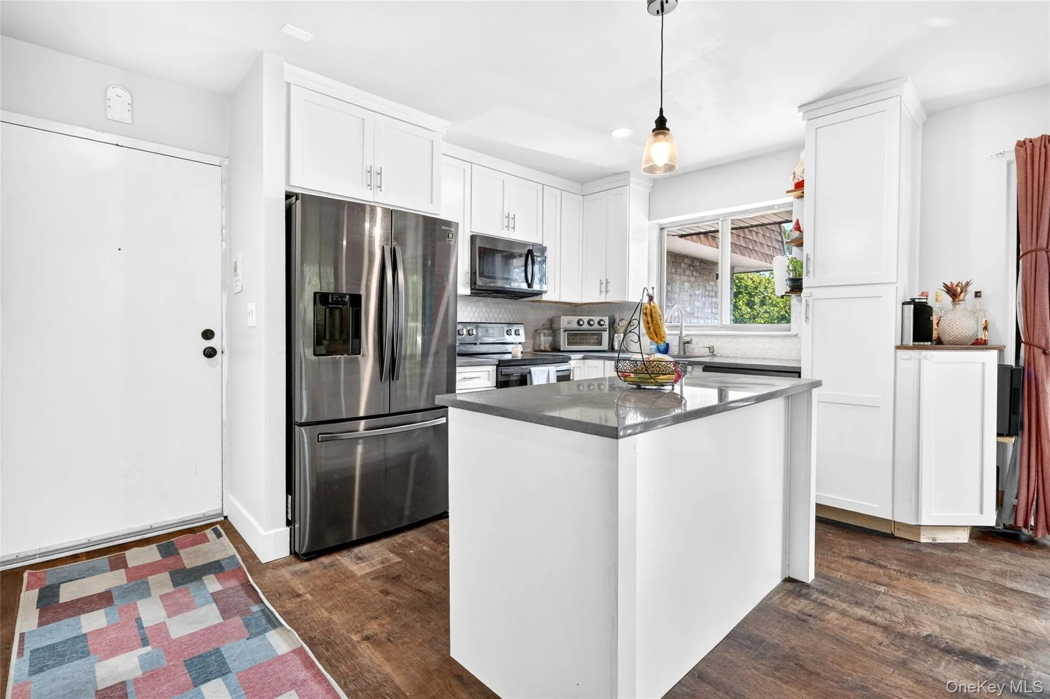 Modern kitchen featuring white cabinetry, stainless steel appliances, a central island with a solid surface countertop, wood-finish flooring, and a subway tile backsplash Modern kitchen featuring white cabinetry, stainless steel appliances, a central island with a solid surface countertop, wood-finish flooring, and a subway tile backsplash