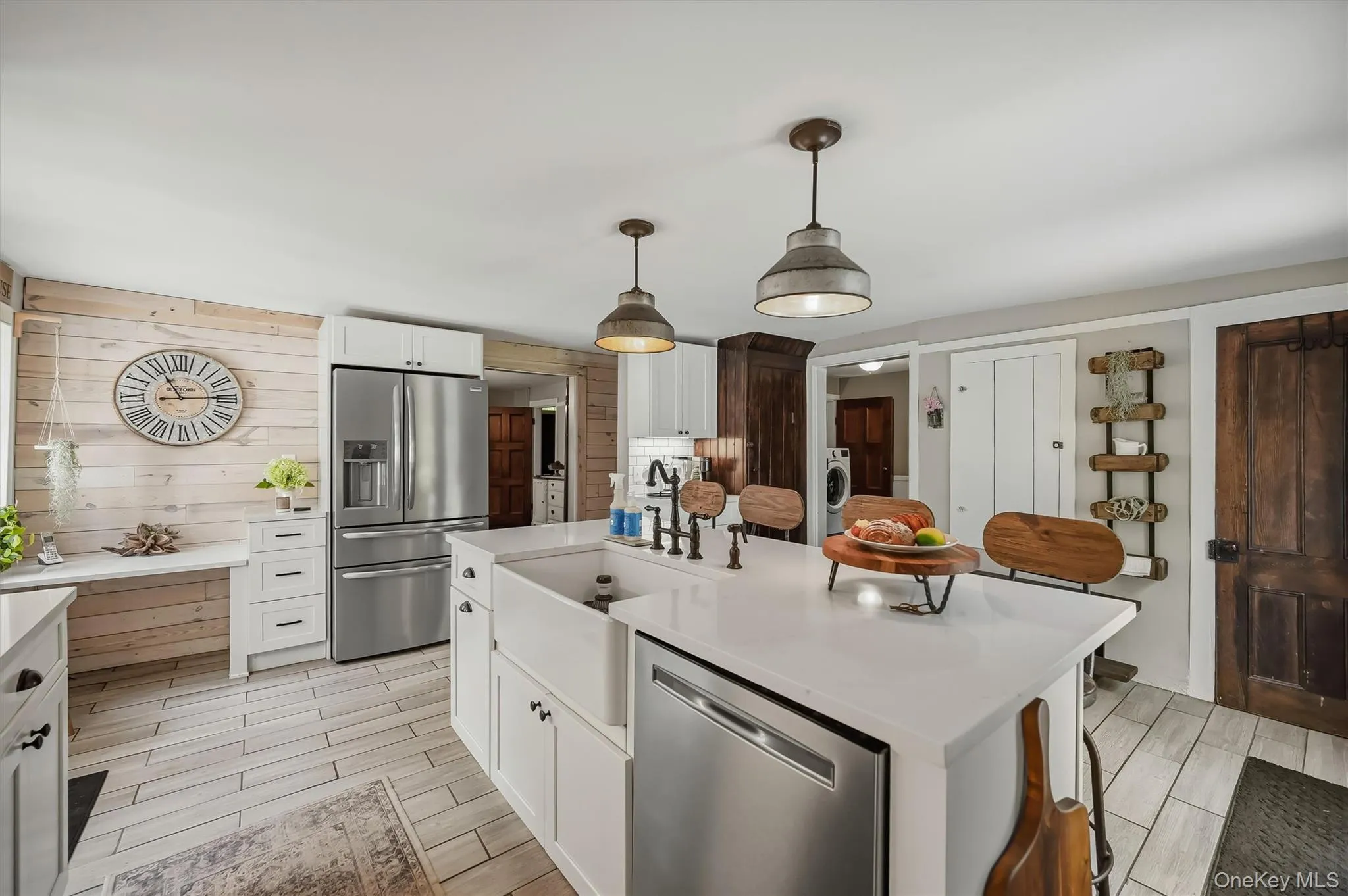 Open-concept kitchen featuring an oversized island with an integrated farmhouse sink, white shaker cabinetry with dark hardware, stainless steel appliances, and wood-finish tile flooring Open-concept kitchen featuring an oversized island with an integrated farmhouse sink, white shaker cabinetry with dark hardware, stainless steel appliances, and wood-finish tile flooring