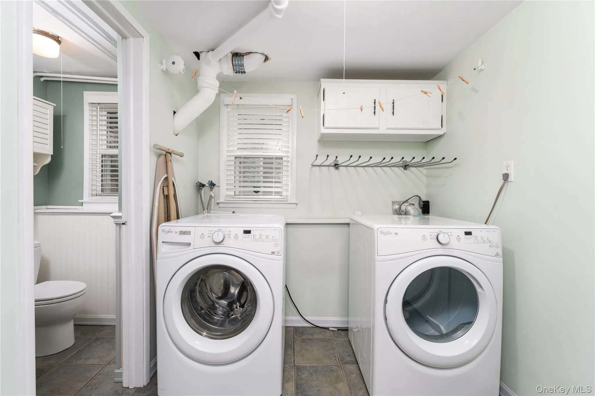 Laundry area featuring two front-loading machines, overhead cabinetry, and a window. Half bath off family room Laundry area featuring two front-loading machines, overhead cabinetry, and a window. Half bath off family room