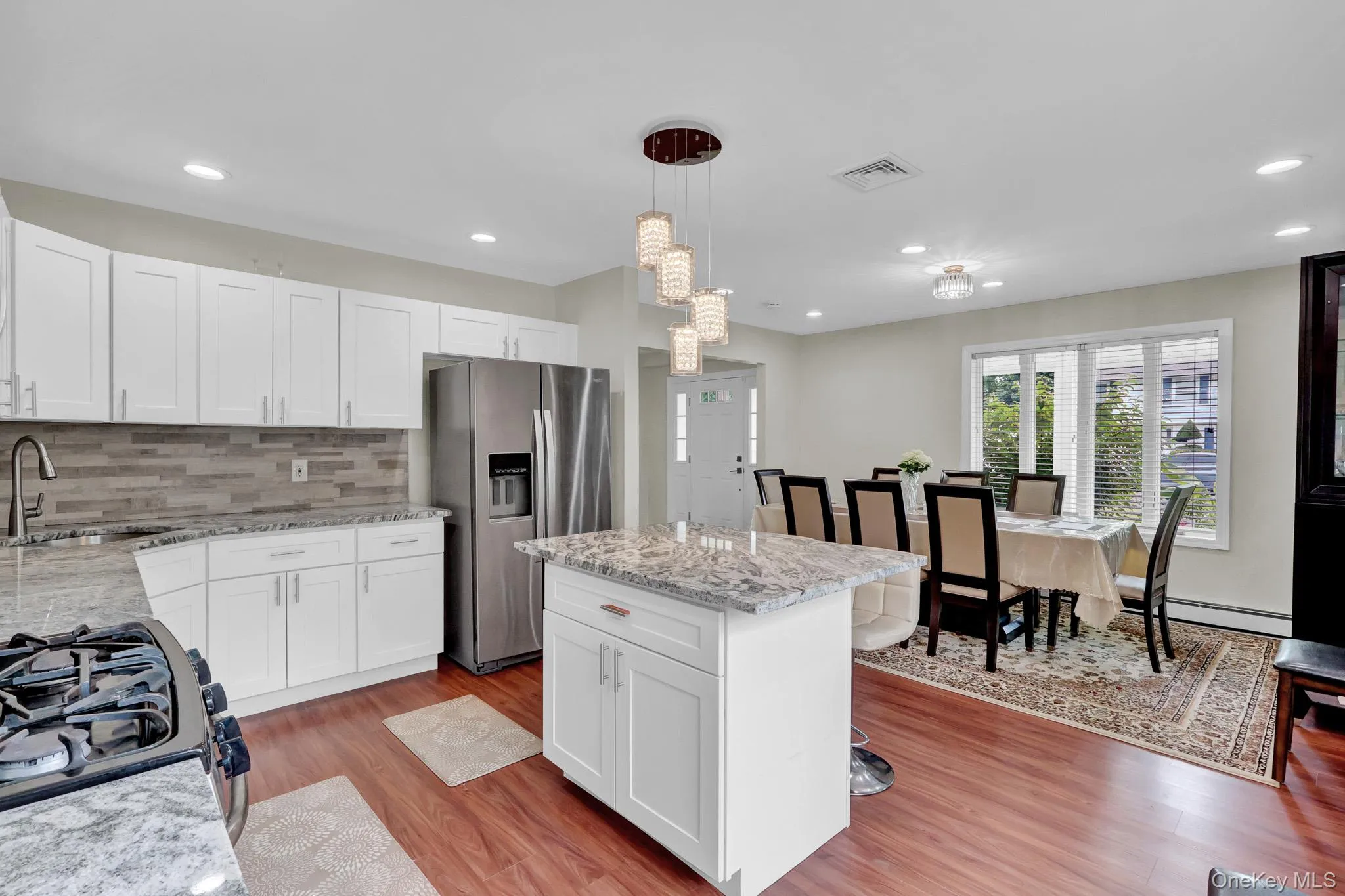 Open-concept kitchen and dining area featuring white shaker-style cabinetry, stainless steel appliances, granite countertops, and wood-finish flooring Open-concept kitchen and dining area featuring white shaker-style cabinetry, stainless steel appliances, granite countertops, and wood-finish flooring