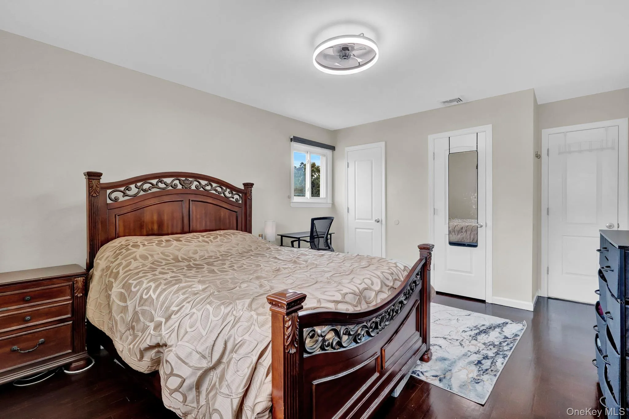 Spacious bedroom featuring dark wood-finish flooring, a contemporary ceiling light fixture, a window with exterior views, and multiple white panel doors Spacious bedroom featuring dark wood-finish flooring, a contemporary ceiling light fixture, a window with exterior views, and multiple white panel doors