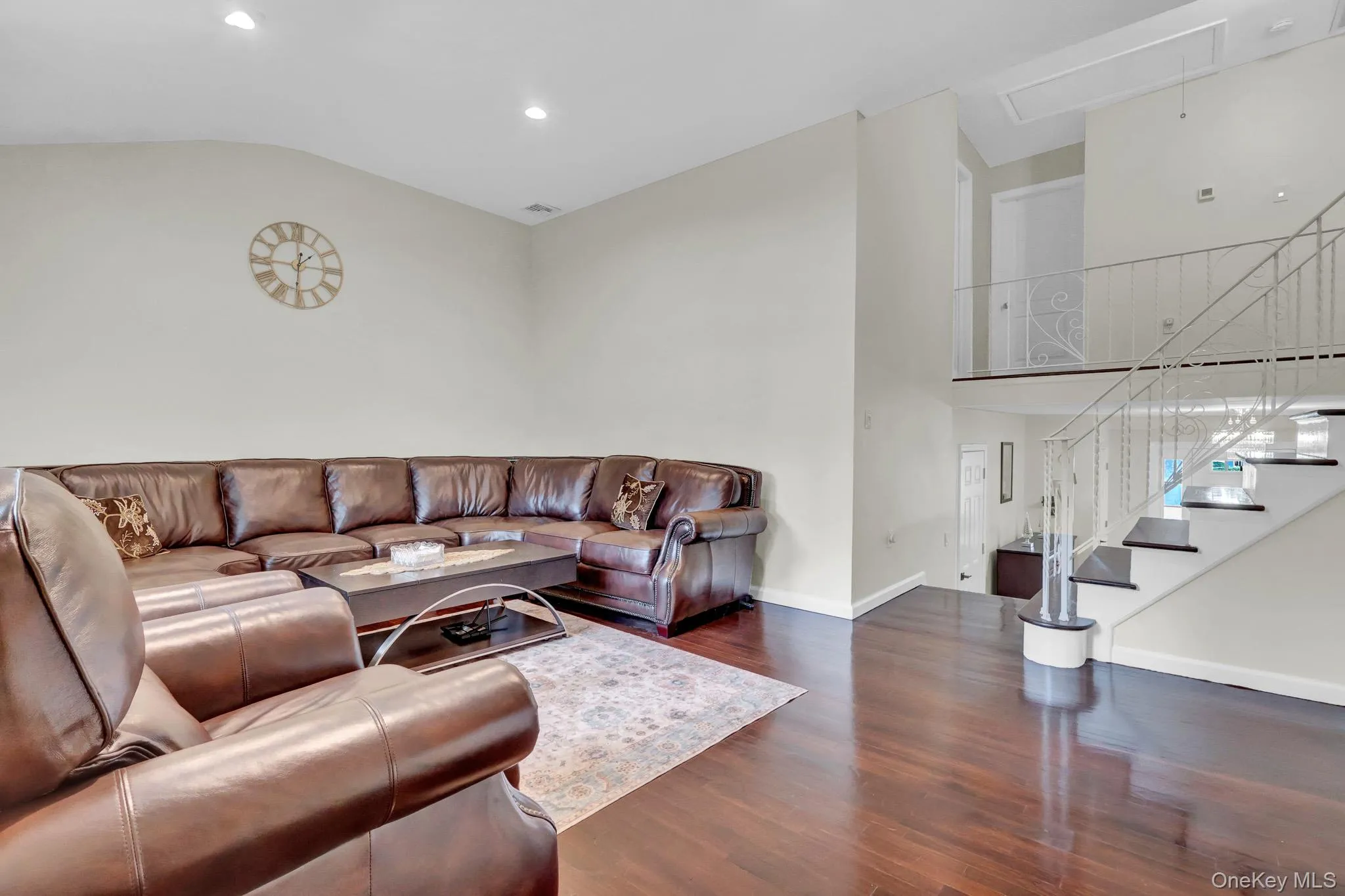 Spacious living area featuring rich wood-finish flooring, recessed lighting, and a prominent open staircase with a decorative metal railing Spacious living area featuring rich wood-finish flooring, recessed lighting, and a prominent open staircase with a decorative metal railing