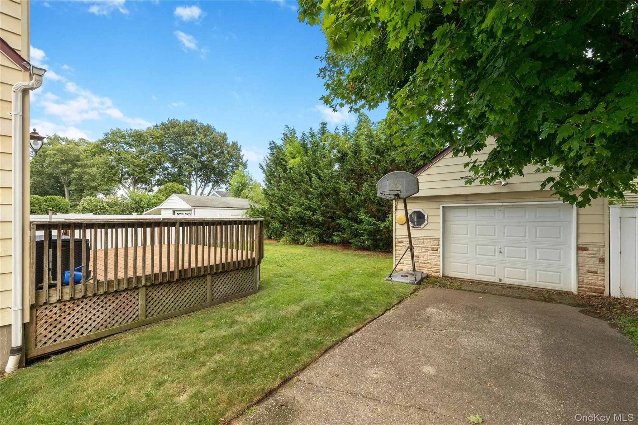 Expansive backyard featuring a wood deck with lattice skirting, a detached garage with a white panel door, and a concrete driveway Expansive backyard featuring a wood deck with lattice skirting, a detached garage with a white panel door, and a concrete driveway