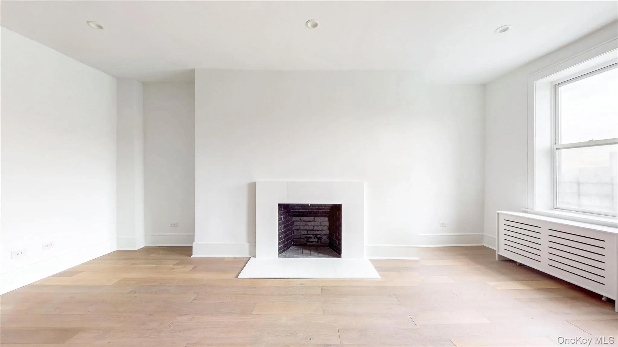 Contemporary living space featuring a white mantel fireplace with a brick firebox, light wood-finish flooring, recessed ceiling lighting, a large window, and a louvered radiator cover Contemporary living space featuring a white mantel fireplace with a brick firebox, light wood-finish flooring, recessed ceiling lighting, a large window, and a louvered radiator cover