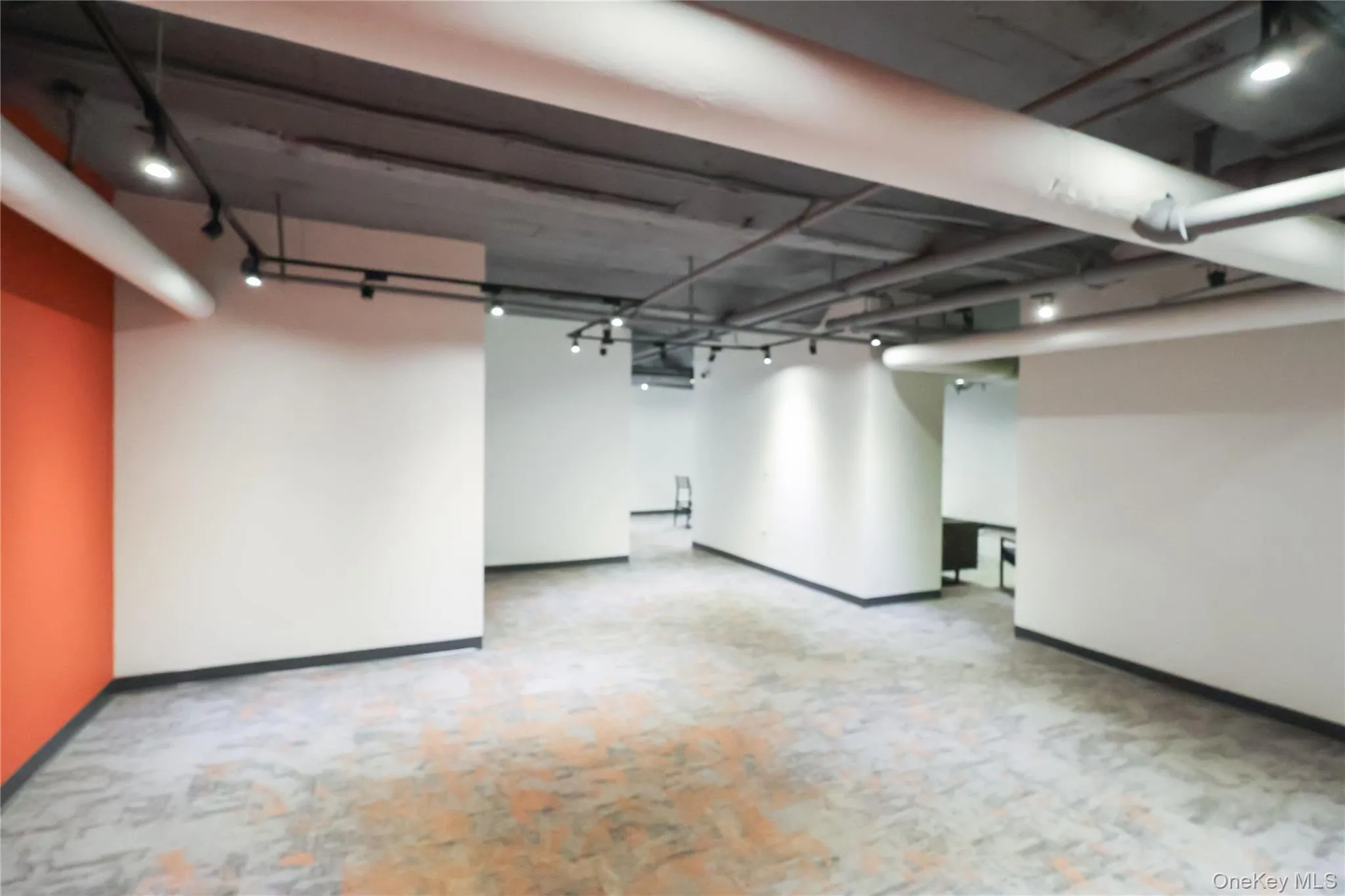 Spacious interior featuring exposed concrete ceilings with track lighting, industrial ductwork, white and orange painted walls, and patterned carpet tile flooring Spacious interior featuring exposed concrete ceilings with track lighting, industrial ductwork, white and orange painted walls, and patterned carpet tile flooring