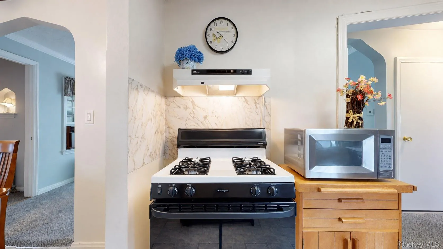 Kitchen area featuring a gas range with an overhead exhaust hood, a marble-patterned backsplash, and a freestanding wood-finish cabinet Kitchen area featuring a gas range with an overhead exhaust hood, a marble-patterned backsplash, and a freestanding wood-finish cabinet
