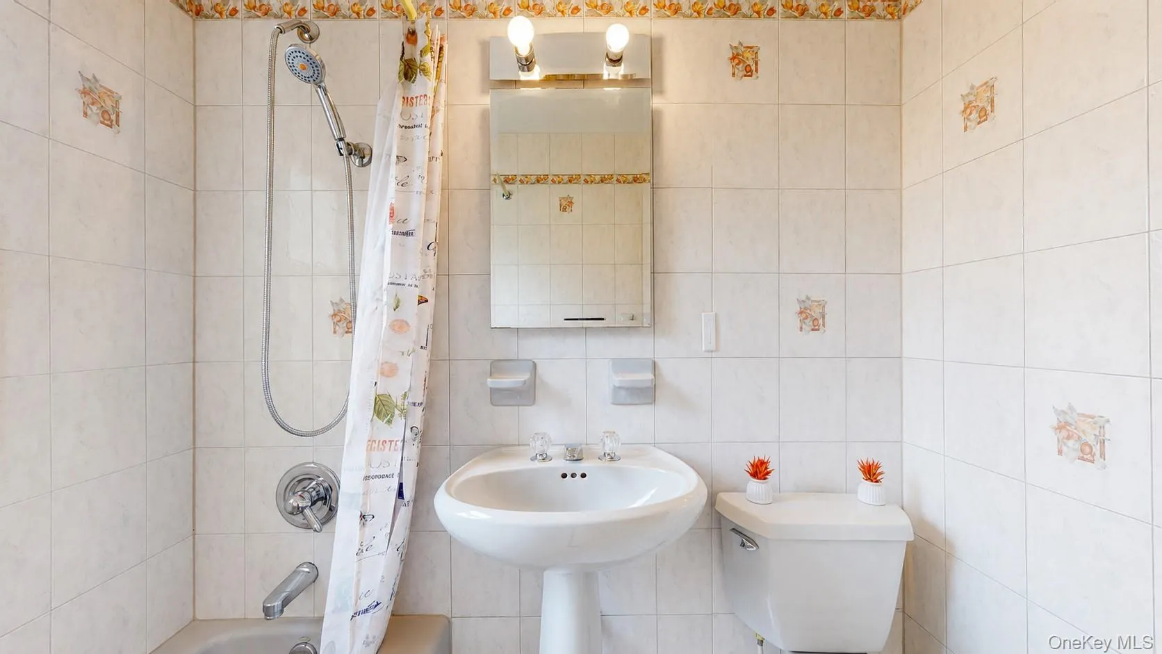Bathroom featuring a pedestal sink, wall-mounted medicine cabinet with integrated lighting, tiled walls with decorative accents, and a tub with a shower fixture Bathroom featuring a pedestal sink, wall-mounted medicine cabinet with integrated lighting, tiled walls with decorative accents, and a tub with a shower fixture