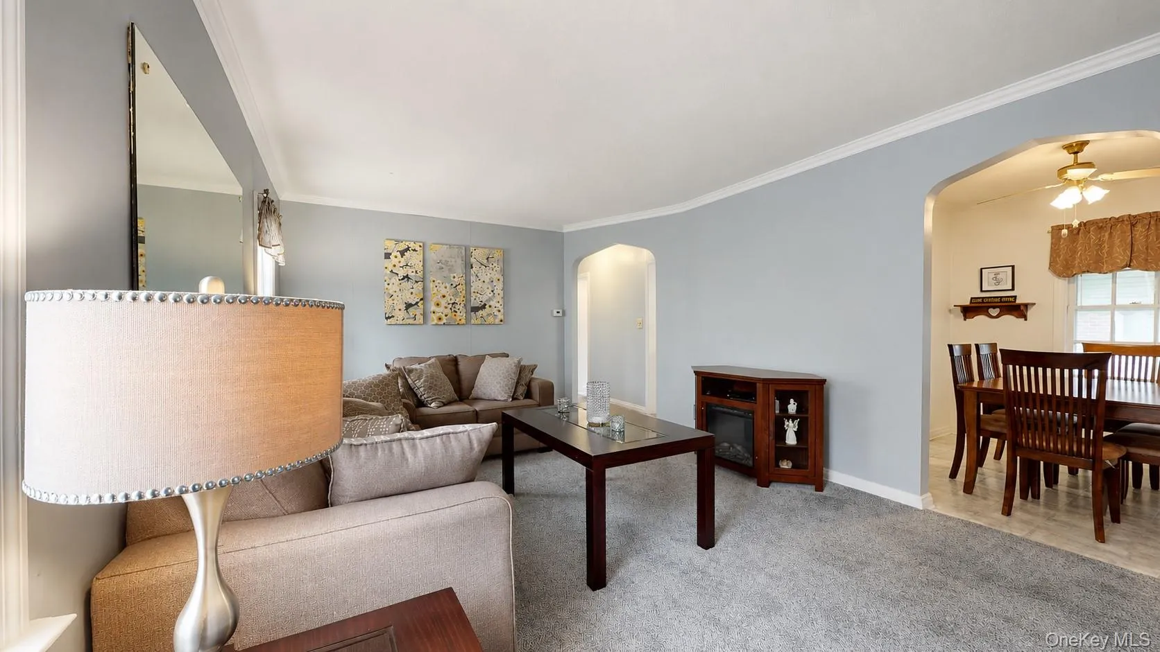 Living room featuring light blue walls, gray carpeting, white crown molding, and an arched doorway opening to a dining area with tiled flooring Living room featuring light blue walls, gray carpeting, white crown molding, and an arched doorway opening to a dining area with tiled flooring