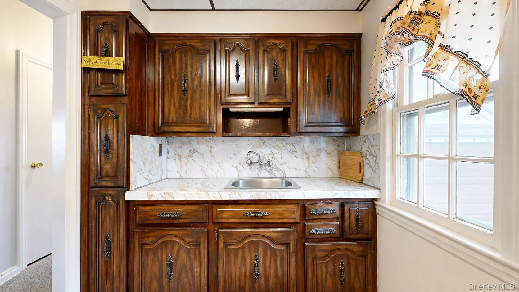 Rich wood cabinetry with decorative hardware, white marble-patterned countertop, stainless steel sink, and a multi-pane window Rich wood cabinetry with decorative hardware, white marble-patterned countertop, stainless steel sink, and a multi-pane window