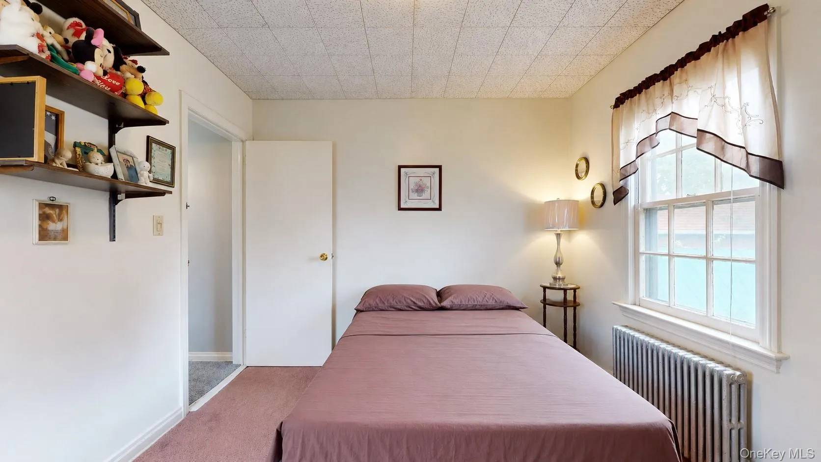 Bedroom featuring a window with a valance, baseboard radiator, wood-finish shelving, and a white interior door Bedroom featuring a window with a valance, baseboard radiator, wood-finish shelving, and a white interior door