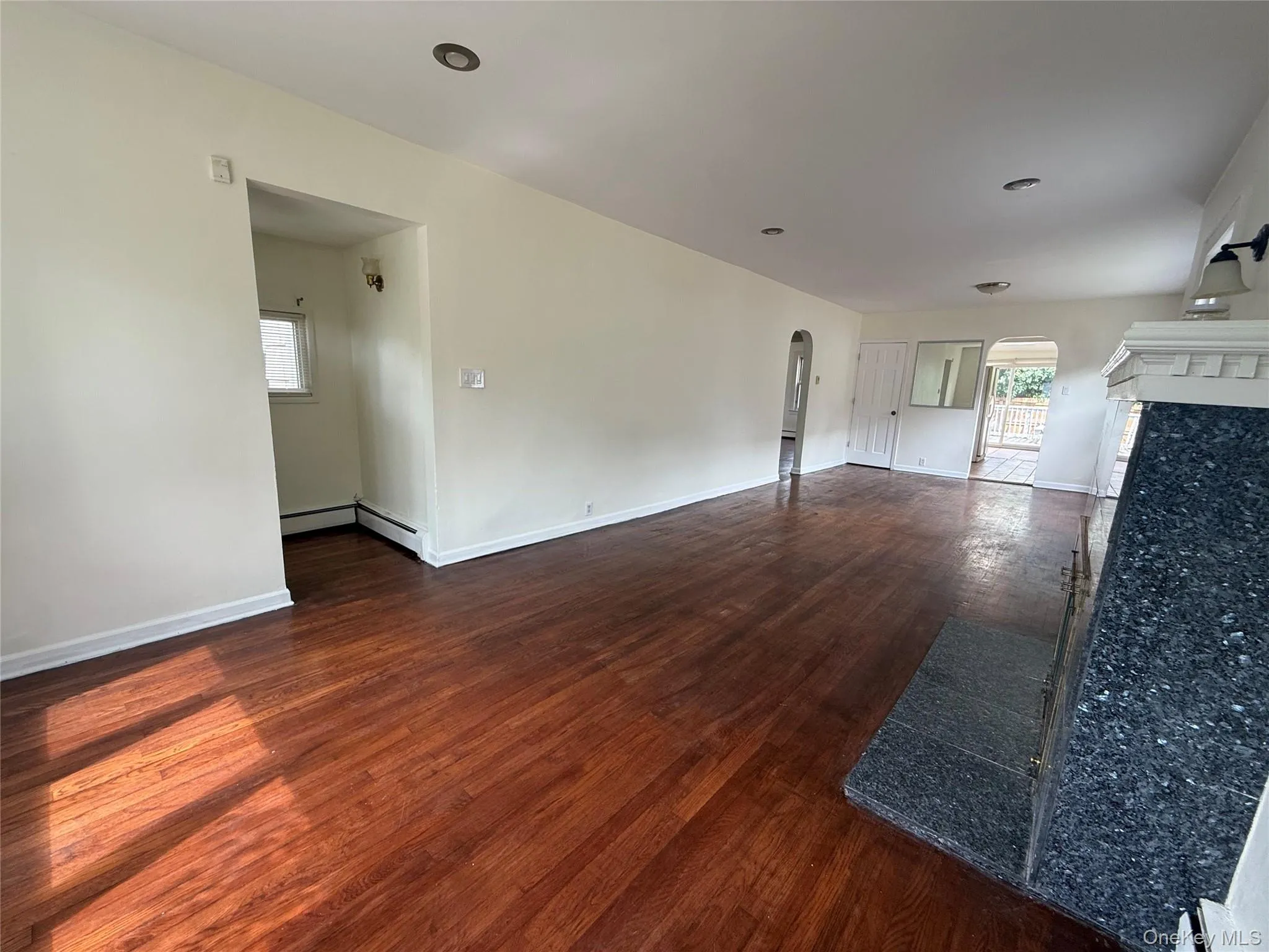 Spacious room featuring wood-finish flooring, light-toned walls, recessed lighting, and a fireplace with a dark, textured stone surround and hearth Spacious room featuring wood-finish flooring, light-toned walls, recessed lighting, and a fireplace with a dark, textured stone surround and hearth