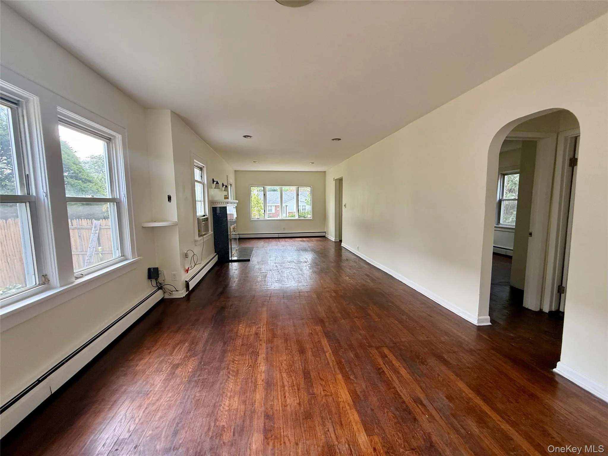 Spacious interior featuring wood-finish flooring, ample natural light from multiple windows, an arched doorway, a fireplace with a dark surround, and baseboard heating Spacious interior featuring wood-finish flooring, ample natural light from multiple windows, an arched doorway, a fireplace with a dark surround, and baseboard heating
