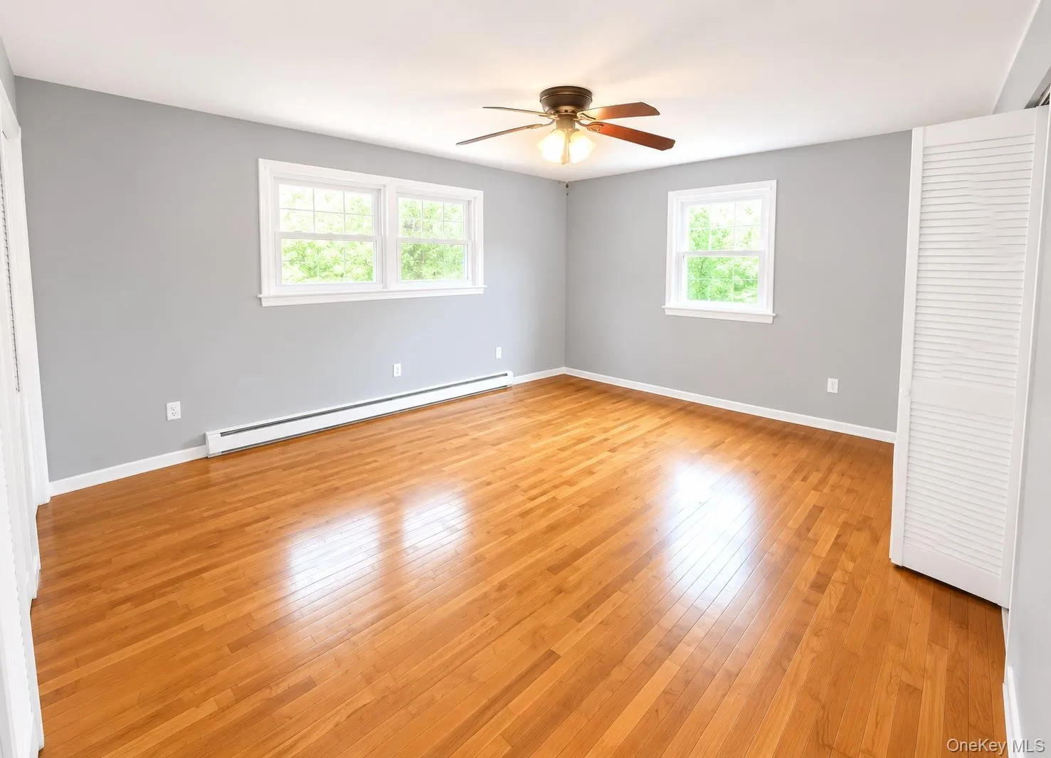 Spacious room featuring wood-finish flooring, two large windows with white trim, a ceiling fan with integrated lighting, and neutral gray wall paint Spacious room featuring wood-finish flooring, two large windows with white trim, a ceiling fan with integrated lighting, and neutral gray wall paint