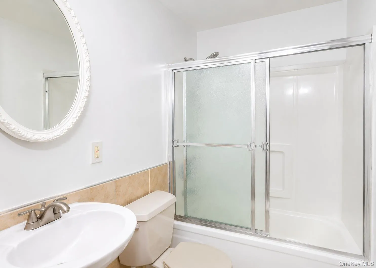 White bathroom featuring a shower-tub combination with sliding glass doors, a pedestal sink with a brushed nickel faucet, a toilet, and a decorative oval mirror White bathroom featuring a shower-tub combination with sliding glass doors, a pedestal sink with a brushed nickel faucet, a toilet, and a decorative oval mirror
