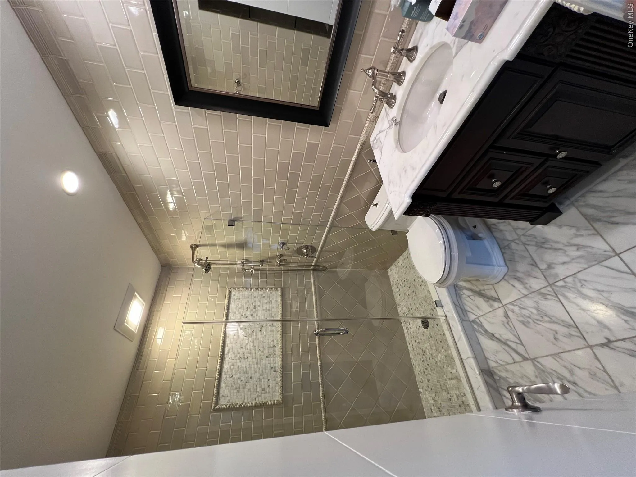 Spa-inspired primary bathroom featuring a frameless glass-enclosed walk-in shower with subway tile surround and pebble stone flooring, dual vanity with white quartz countertop and matte black hardware, framed mirror, and marble-look floor tile throughout Spa-inspired primary bathroom featuring a frameless glass-enclosed walk-in shower with subway tile surround and pebble stone flooring, dual vanity with white quartz countertop and matte black hardware, framed mirror, and marble-look floor tile throughout