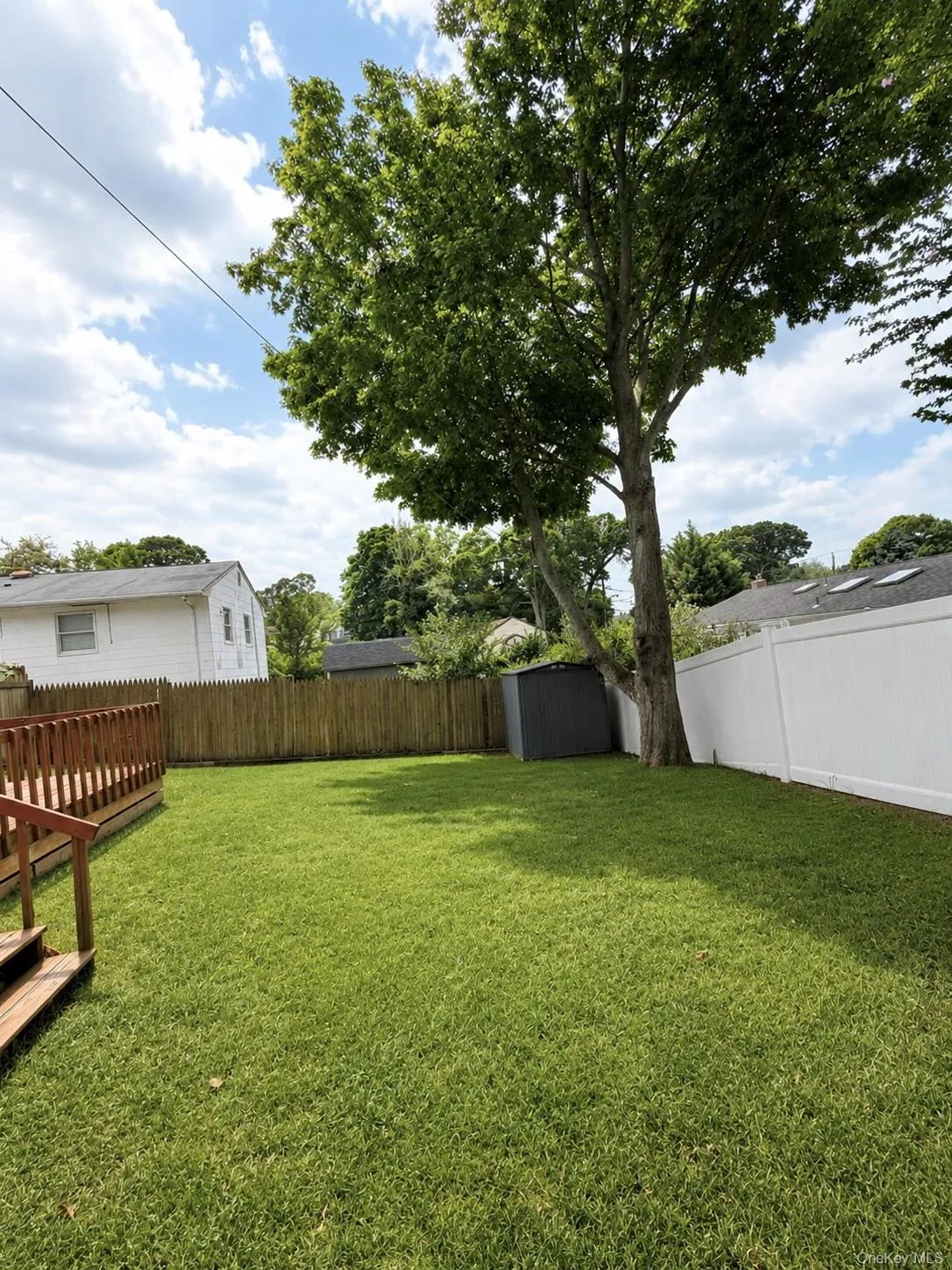 Green lawn, a wooden deck, a privacy fence along the perimeter, and mature trees providing natural shade. Green lawn, a wooden deck, a privacy fence along the perimeter, and mature trees providing natural shade.