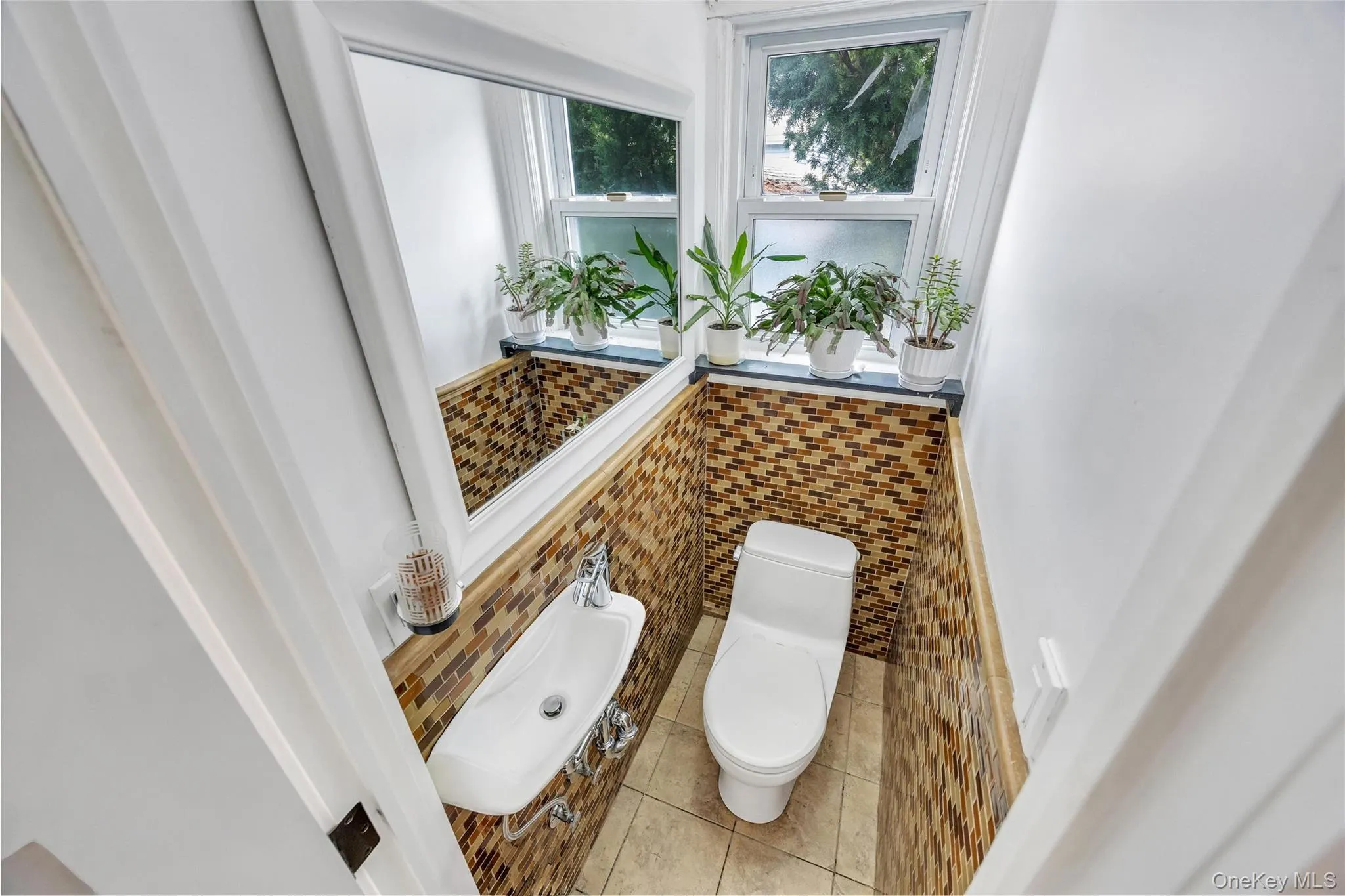 Bathroom featuring mosaic tile wainscoting, a wall-mounted sink with chrome fixtures, a modern toilet, and an overhead window Bathroom featuring mosaic tile wainscoting, a wall-mounted sink with chrome fixtures, a modern toilet, and an overhead window