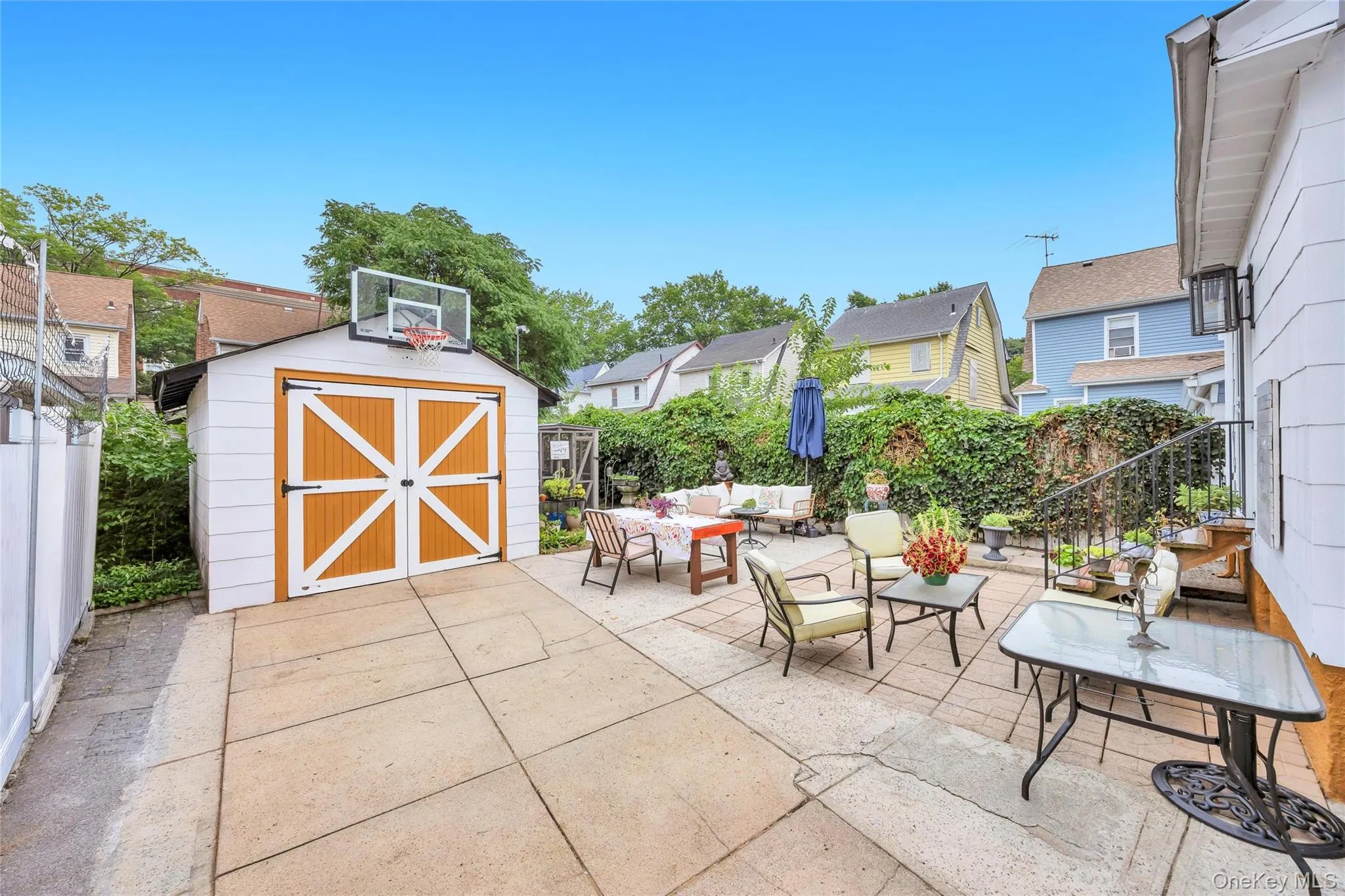 Expansive concrete patio area featuring a detached shed with double doors, established perimeter fencing, and mature greenery Expansive concrete patio area featuring a detached shed with double doors, established perimeter fencing, and mature greenery