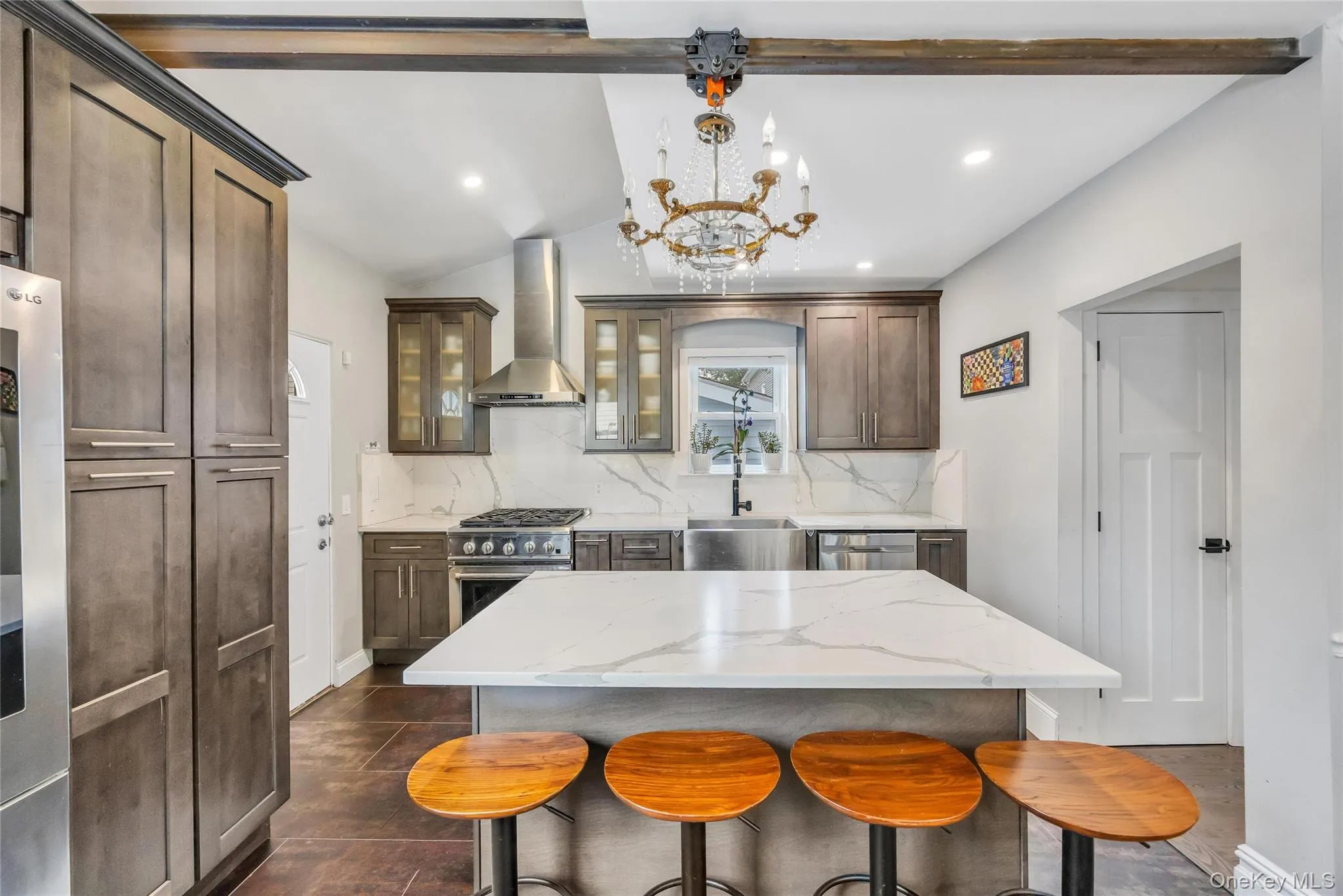 Contemporary kitchen featuring a large central island with a solid surface countertop, stainless steel appliances, dark wood cabinetry, and a decorative chandelier Contemporary kitchen featuring a large central island with a solid surface countertop, stainless steel appliances, dark wood cabinetry, and a decorative chandelier
