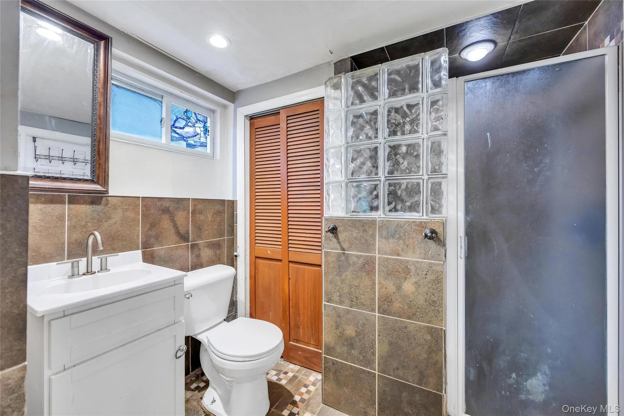 Bathroom featuring a white vanity with a single basin sink, a toilet, a glass block shower enclosure, and a frosted shower door Bathroom featuring a white vanity with a single basin sink, a toilet, a glass block shower enclosure, and a frosted shower door