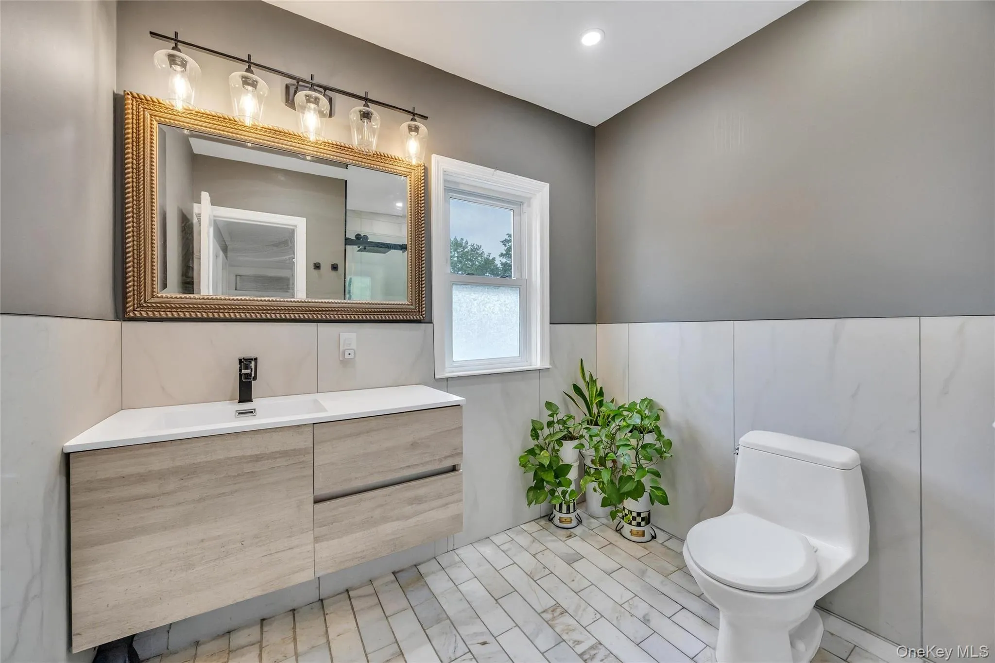 Contemporary bathroom featuring a floating vanity with integrated sink, patterned gold-tone framed mirror, four-light vanity fixture, and floor-to-ceiling white tiled walls with a light gray accent wall Contemporary bathroom featuring a floating vanity with integrated sink, patterned gold-tone framed mirror, four-light vanity fixture, and floor-to-ceiling white tiled walls with a light gray accent wall