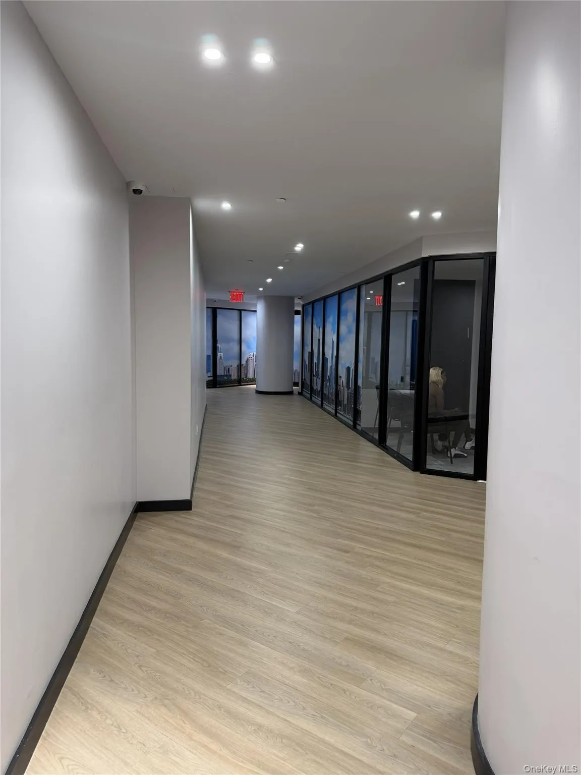Modern hallway featuring light wood-finish flooring, recessed ceiling lighting, white walls, and a curved glass wall partition with black framing Modern hallway featuring light wood-finish flooring, recessed ceiling lighting, white walls, and a curved glass wall partition with black framing