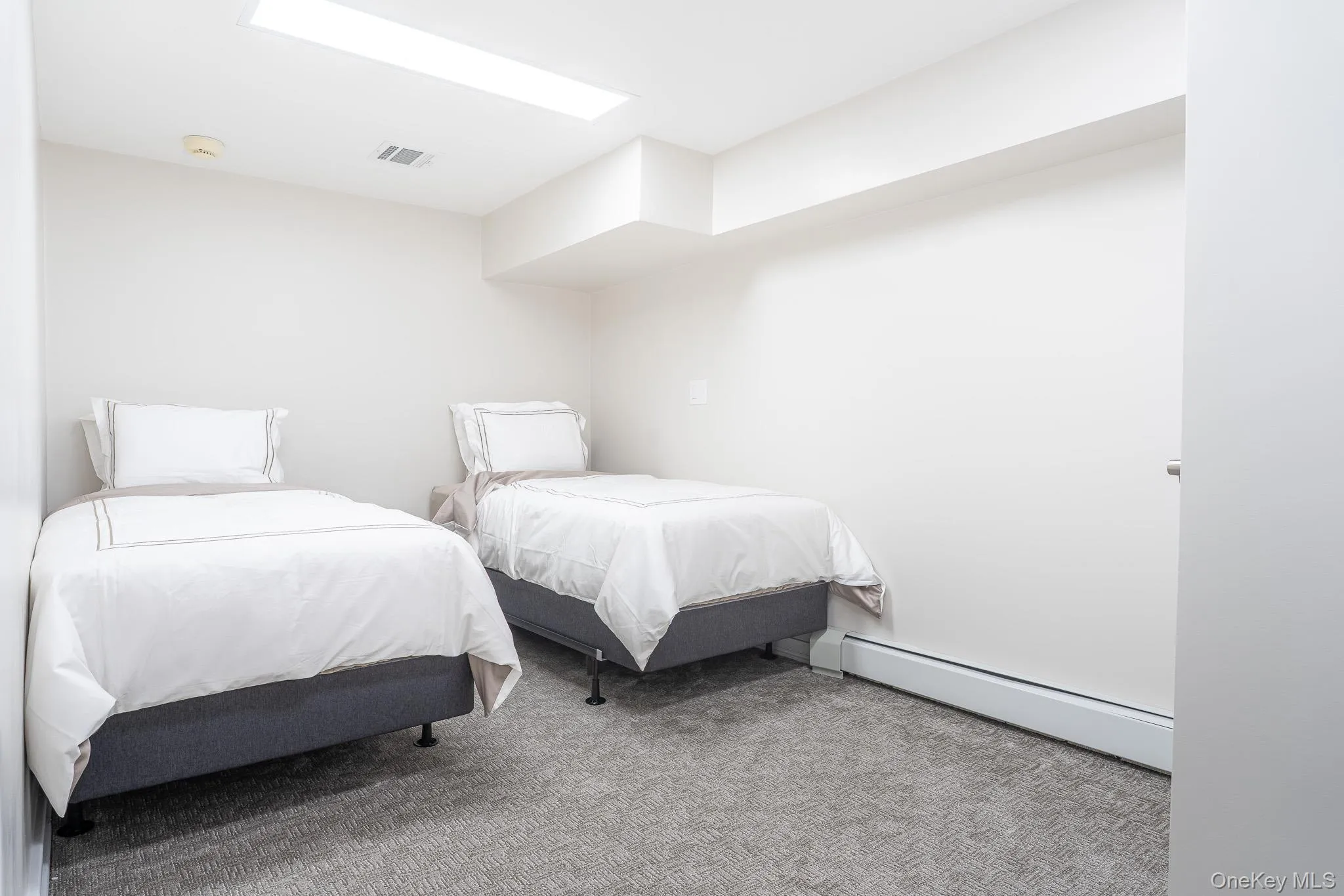 Dual bedroom space featuring two twin beds with white linens and dark base platforms, gray carpet flooring, recessed ceiling lighting, and clean white walls Dual bedroom space featuring two twin beds with white linens and dark base platforms, gray carpet flooring, recessed ceiling lighting, and clean white walls