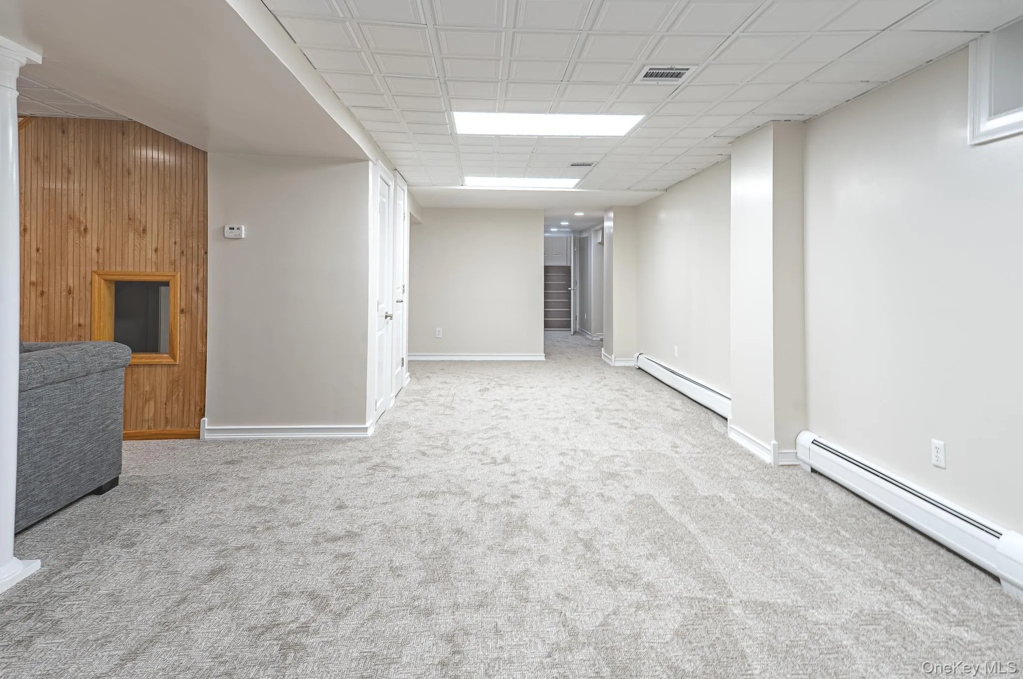 Spacious carpeted room featuring a drop ceiling with recessed lighting, baseboard heating, white painted walls, and an accent wall with vertical wood paneling and a framed opening Spacious carpeted room featuring a drop ceiling with recessed lighting, baseboard heating, white painted walls, and an accent wall with vertical wood paneling and a framed opening