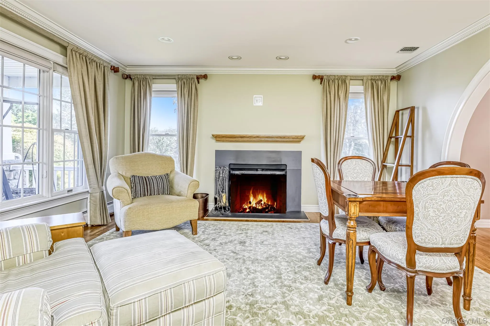 Elegant living room featuring a wood-burning fireplace with surround mantel, large multi-pane windows with custom drapery, refinished wood-frame seating, and neutral plush area rug Elegant living room featuring a wood-burning fireplace with surround mantel, large multi-pane windows with custom drapery, refinished wood-frame seating, and neutral plush area rug