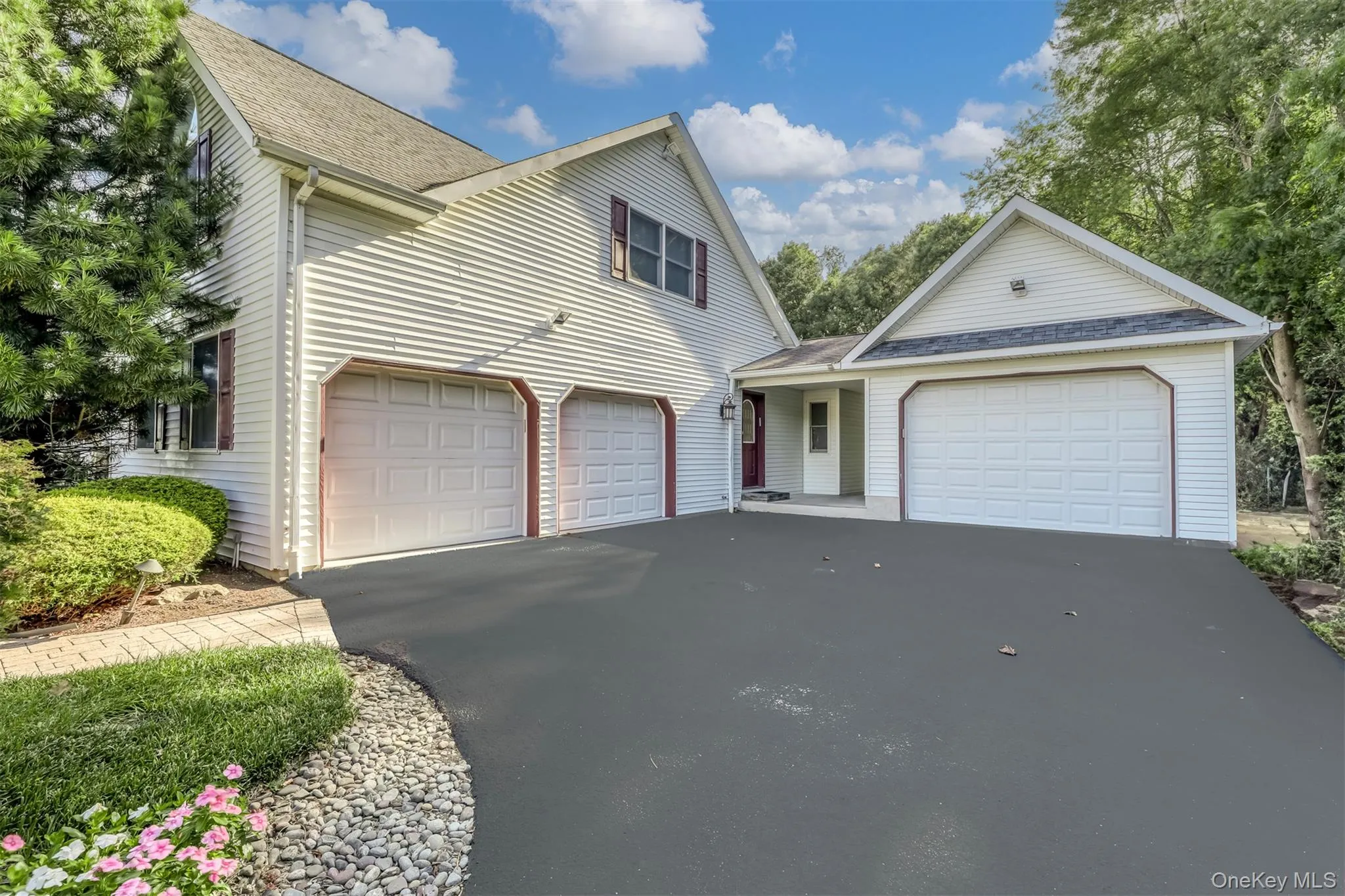 Freshly sealed asphalt driveway anchors this two-garage home featuring white vinyl siding, a peaked roofline with architectural shingles, and manicured landscaping with mature trees and flowering shrubs Freshly sealed asphalt driveway anchors this two-garage home featuring white vinyl siding, a peaked roofline with architectural shingles, and manicured landscaping with mature trees and flowering shrubs