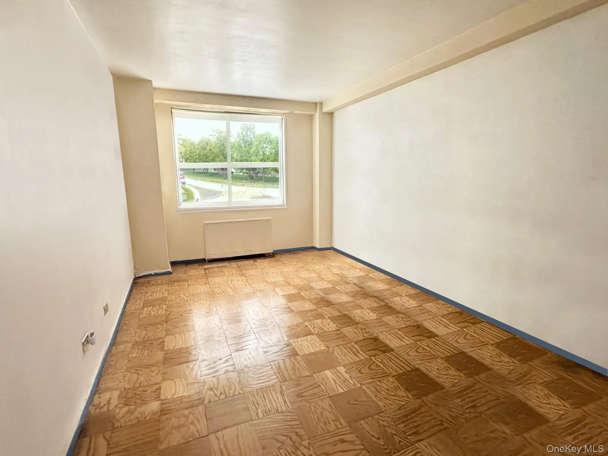 Room featuring parquet wood-finish flooring, a large window, a wall-mounted radiator, and light-toned walls Room featuring parquet wood-finish flooring, a large window, a wall-mounted radiator, and light-toned walls