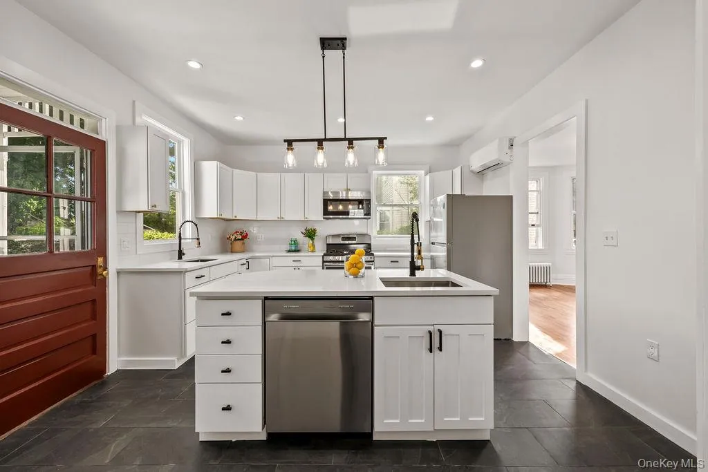 Modern kitchen featuring a central island with an undermount sink, white cabinetry with black hardware, stainless steel appliances, and dark grey tile flooring Modern kitchen featuring a central island with an undermount sink, white cabinetry with black hardware, stainless steel appliances, and dark grey tile flooring