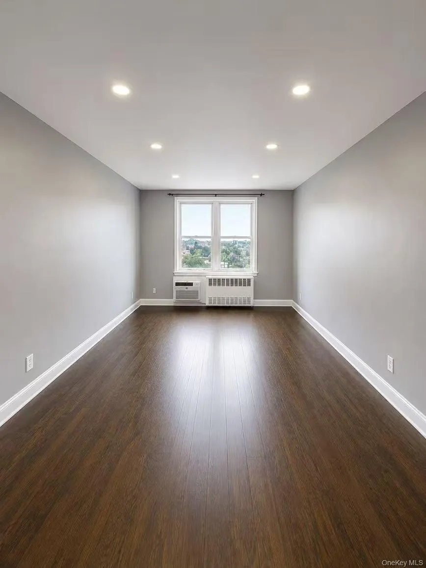 Spacious room featuring wood-finish flooring, recessed lighting, and a double window with a window-mounted air conditioning unit and radiator below Spacious room featuring wood-finish flooring, recessed lighting, and a double window with a window-mounted air conditioning unit and radiator below