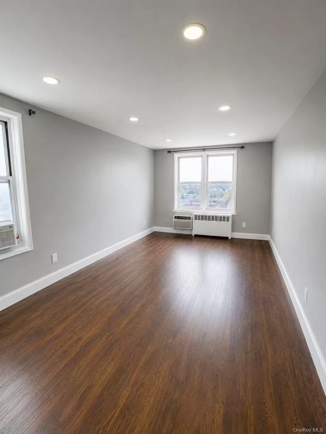 Spacious room featuring wood-finish flooring, light gray walls, white baseboards, recessed lighting, and multiple windows Spacious room featuring wood-finish flooring, light gray walls, white baseboards, recessed lighting, and multiple windows