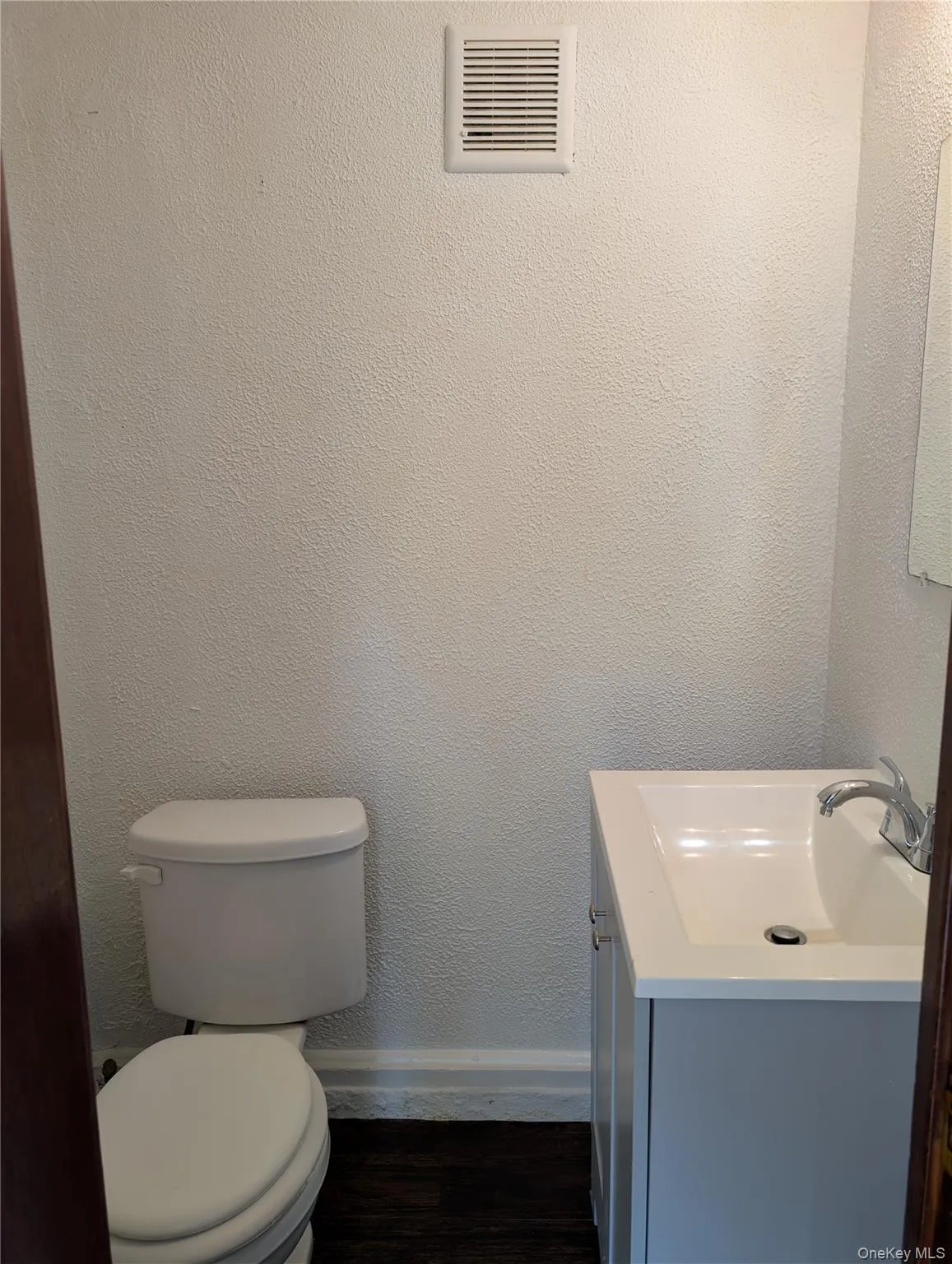 Half bath with white ceramic toilet, gray vanity with integrated sink, dark wood-finish flooring, and white wall finishes Half bath with white ceramic toilet, gray vanity with integrated sink, dark wood-finish flooring, and white wall finishes