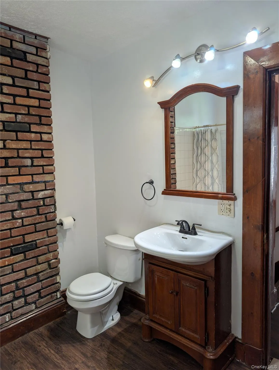 Bathroom featuring a brick-finish wall, vinyl wood-finish flooring, a vanity with a white ceramic sink, and a decorative wood-framed mirror Bathroom featuring a brick-finish wall, vinyl wood-finish flooring, a vanity with a white ceramic sink, and a decorative wood-framed mirror