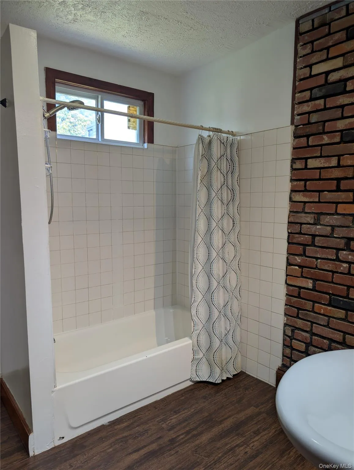 Updated bathroom featuring a white surround, with chrome hardware, white bathtub with integrated apron, vinyl wood-finish flooring, and an exposed brick accent wall with large transom window providing natural light Updated bathroom featuring a white surround, with chrome hardware, white bathtub with integrated apron, vinyl wood-finish flooring, and an exposed brick accent wall with large transom window providing natural light