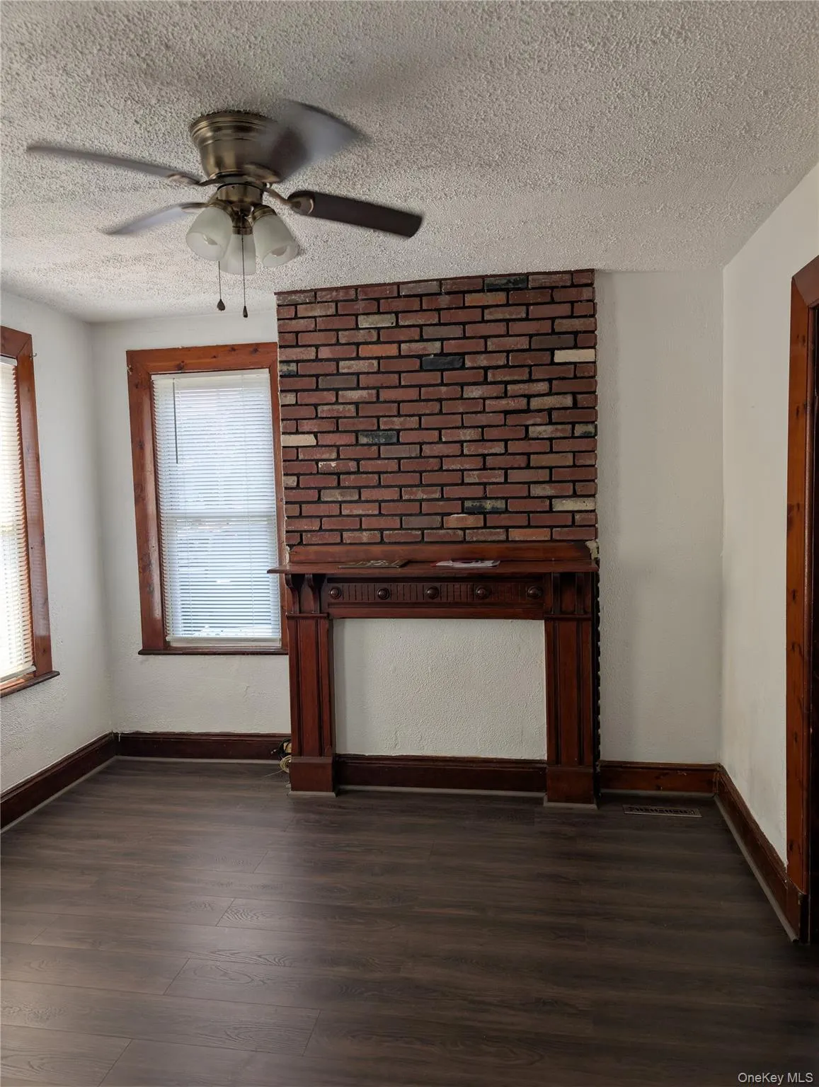 Spacious living room featuring vinyl flooring, an exposed brick hearth with wood mantel, ceiling fan with pull-chain controls, and large windows with wooden frames allowing natural light Spacious living room featuring vinyl flooring, an exposed brick hearth with wood mantel, ceiling fan with pull-chain controls, and large windows with wooden frames allowing natural light