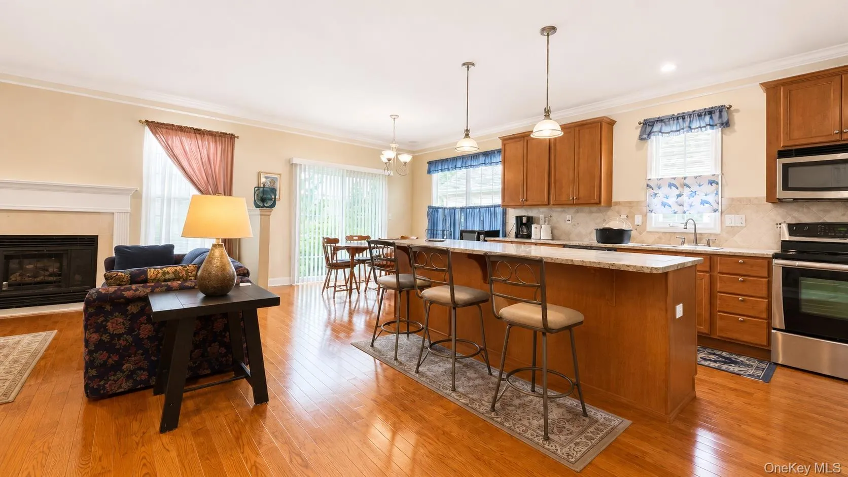 Hardwood flooring throughout, a fireplace with a white mantel, a kitchen island with seating, pendant lighting, and stainless steel appliances Hardwood flooring throughout, a fireplace with a white mantel, a kitchen island with seating, pendant lighting, and stainless steel appliances