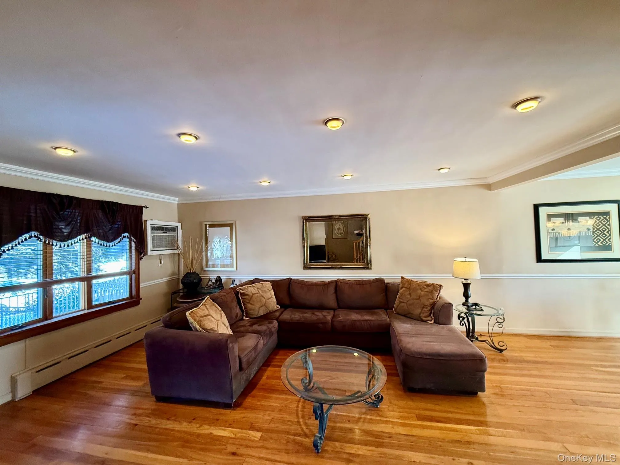 Spacious living area featuring wood-finish flooring, recessed ceiling lighting, crown molding, and a large window with a valance Spacious living area featuring wood-finish flooring, recessed ceiling lighting, crown molding, and a large window with a valance