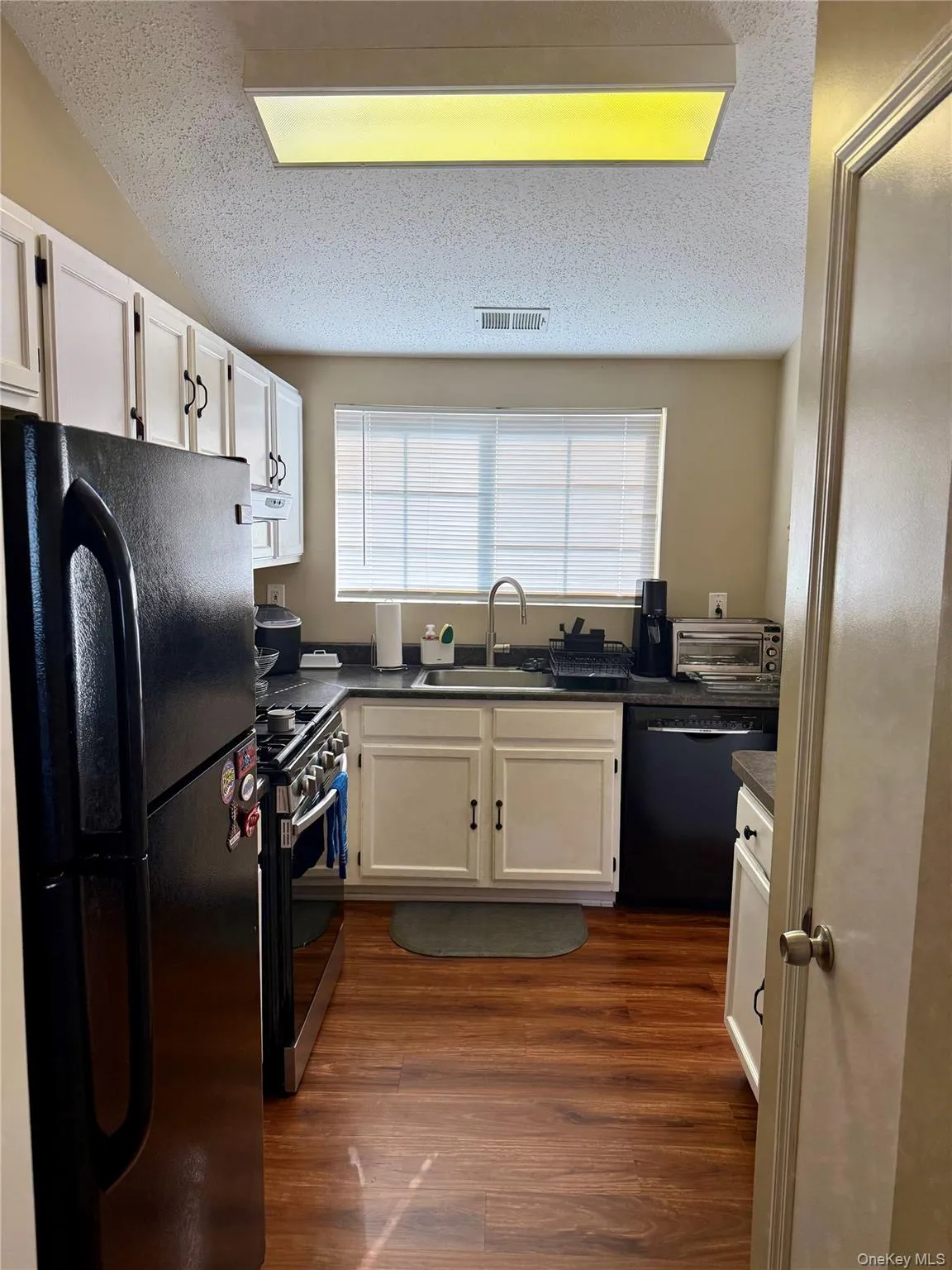 Kitchen with black appliances, a textured ceiling, white cabinets, dark wood-type flooring, and dark countertops Kitchen with black appliances, a textured ceiling, white cabinets, dark wood-type flooring, and dark countertops