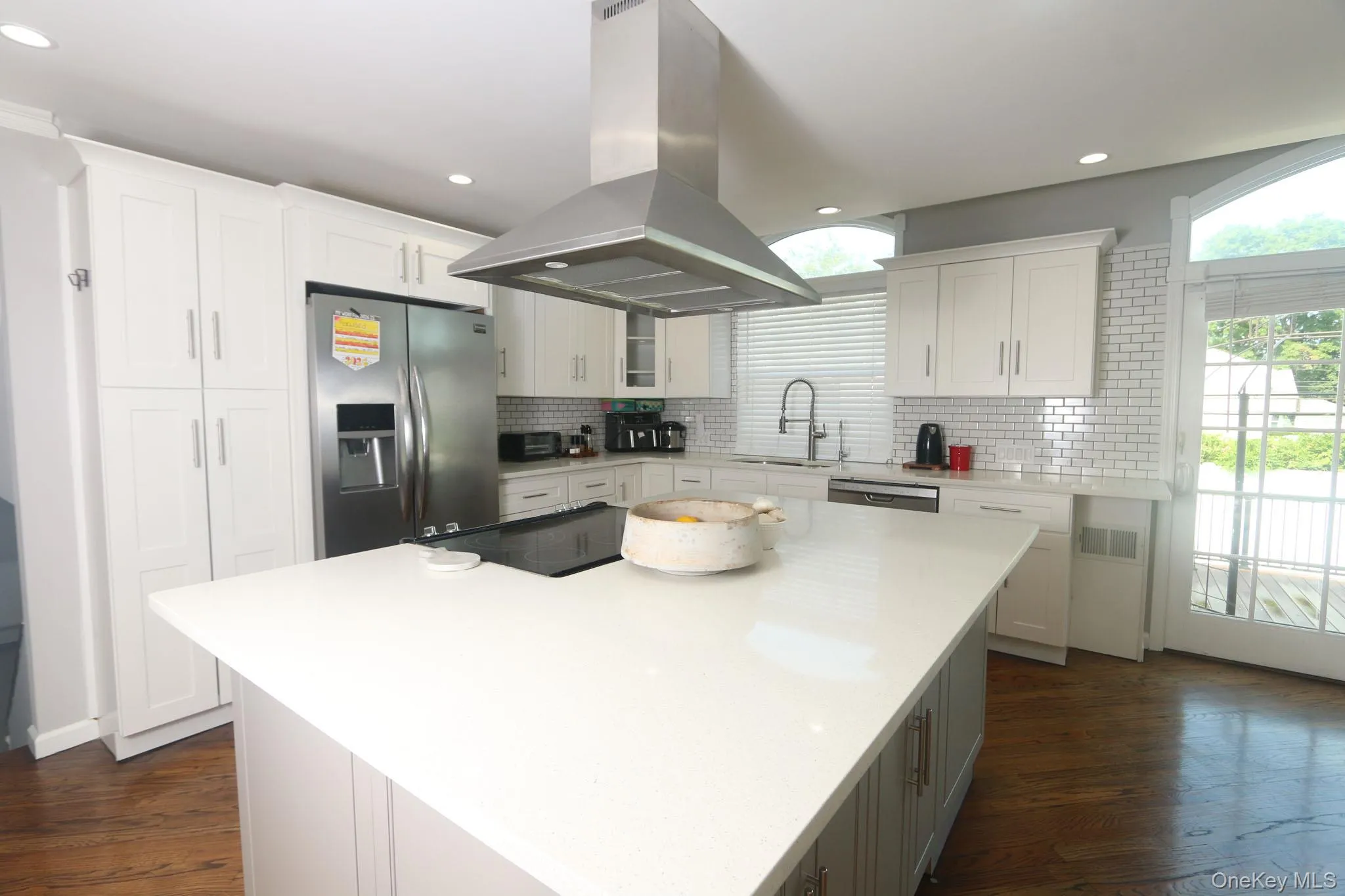 Spacious kitchen featuring an expansive center island, white cabinetry, a stainless steel refrigerator, a professional-grade range hood, and wood-finish flooring Spacious kitchen featuring an expansive center island, white cabinetry, a stainless steel refrigerator, a professional-grade range hood, and wood-finish flooring