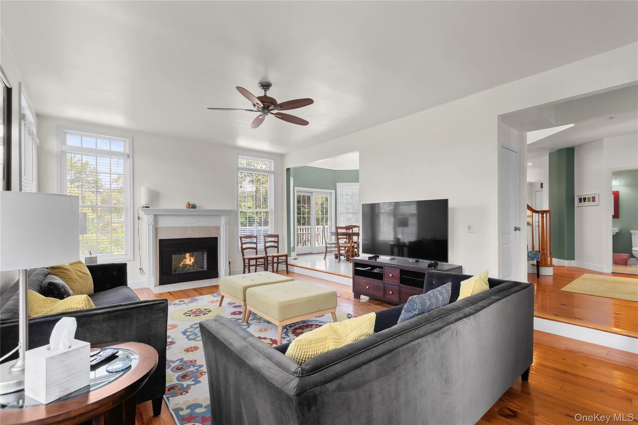 Living area featuring hardwood flooring, a fireplace with a stone and mantel, multiple windows, and a ceiling fan Living area featuring hardwood flooring, a fireplace with a stone and mantel, multiple windows, and a ceiling fan
