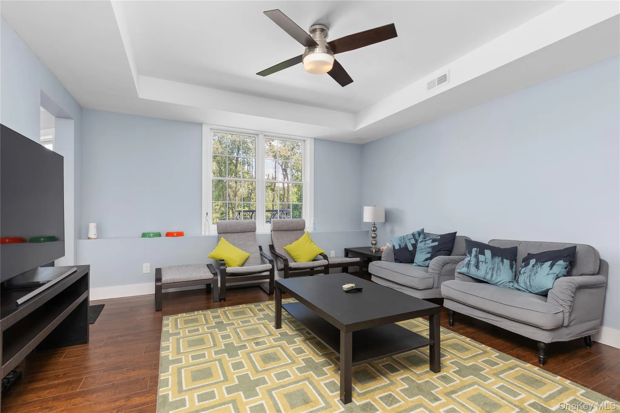 Living area featuring hard wood flooring, a tray ceiling with a ceiling fan, and large windows. Living area featuring hard wood flooring, a tray ceiling with a ceiling fan, and large windows.
