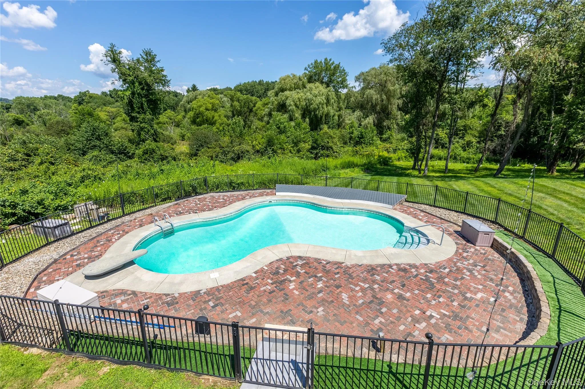 Freeform swimming pool with a brick sitting and lounge area, enclosed by black metal fencing, set against a backdrop of mature trees and green landscaping Freeform swimming pool with a brick sitting and lounge area, enclosed by black metal fencing, set against a backdrop of mature trees and green landscaping