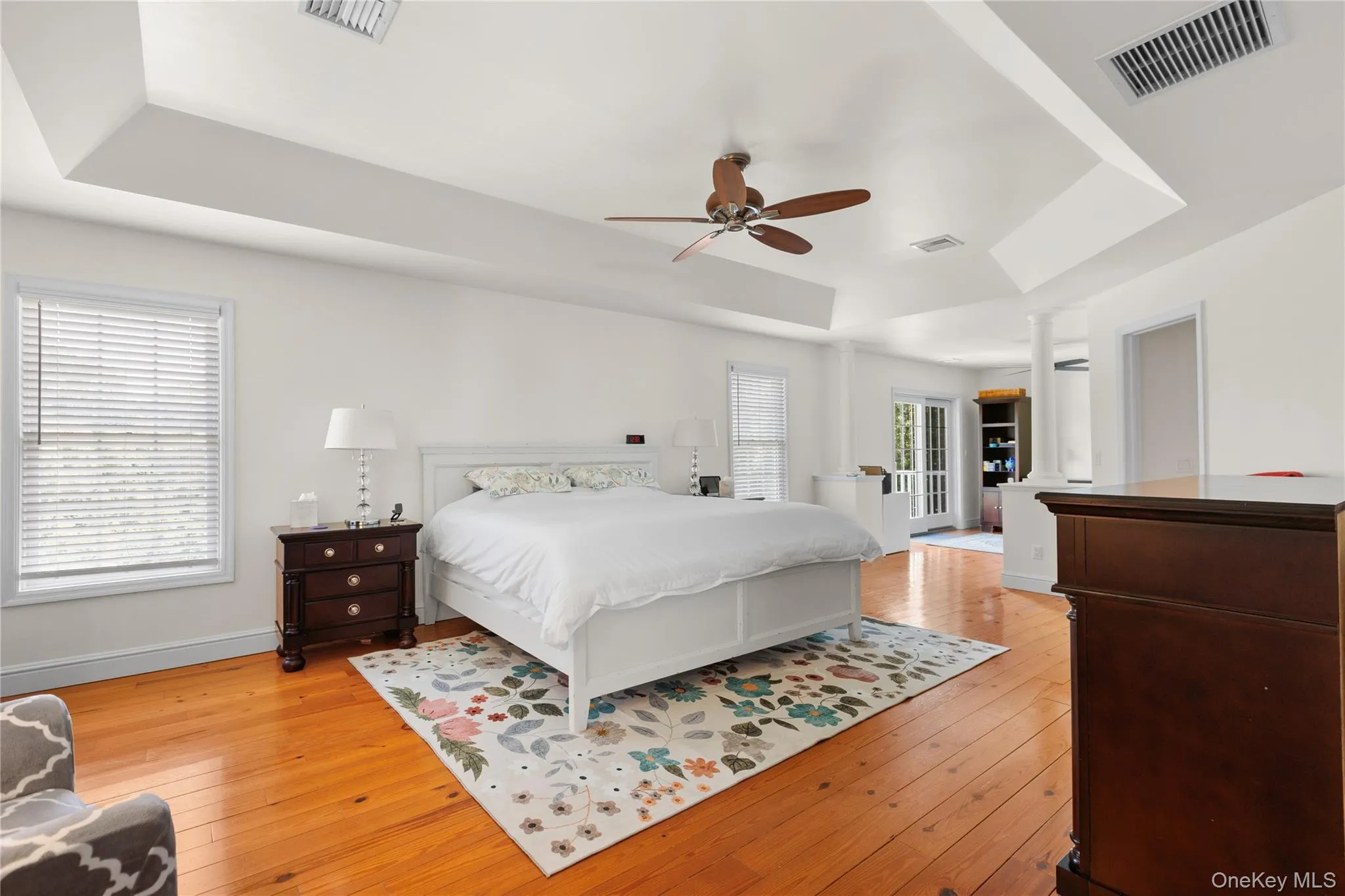 Spacious primary bedroom showcasing coffered ceiling architecture, sitting room with door to balcony, ceiling fan with wood blades, hardwood flooring throughout, and abundant natural light from large windows with plantation shutters Spacious primary bedroom showcasing coffered ceiling architecture, sitting room with door to balcony, ceiling fan with wood blades, hardwood flooring throughout, and abundant natural light from large windows with plantation shutters