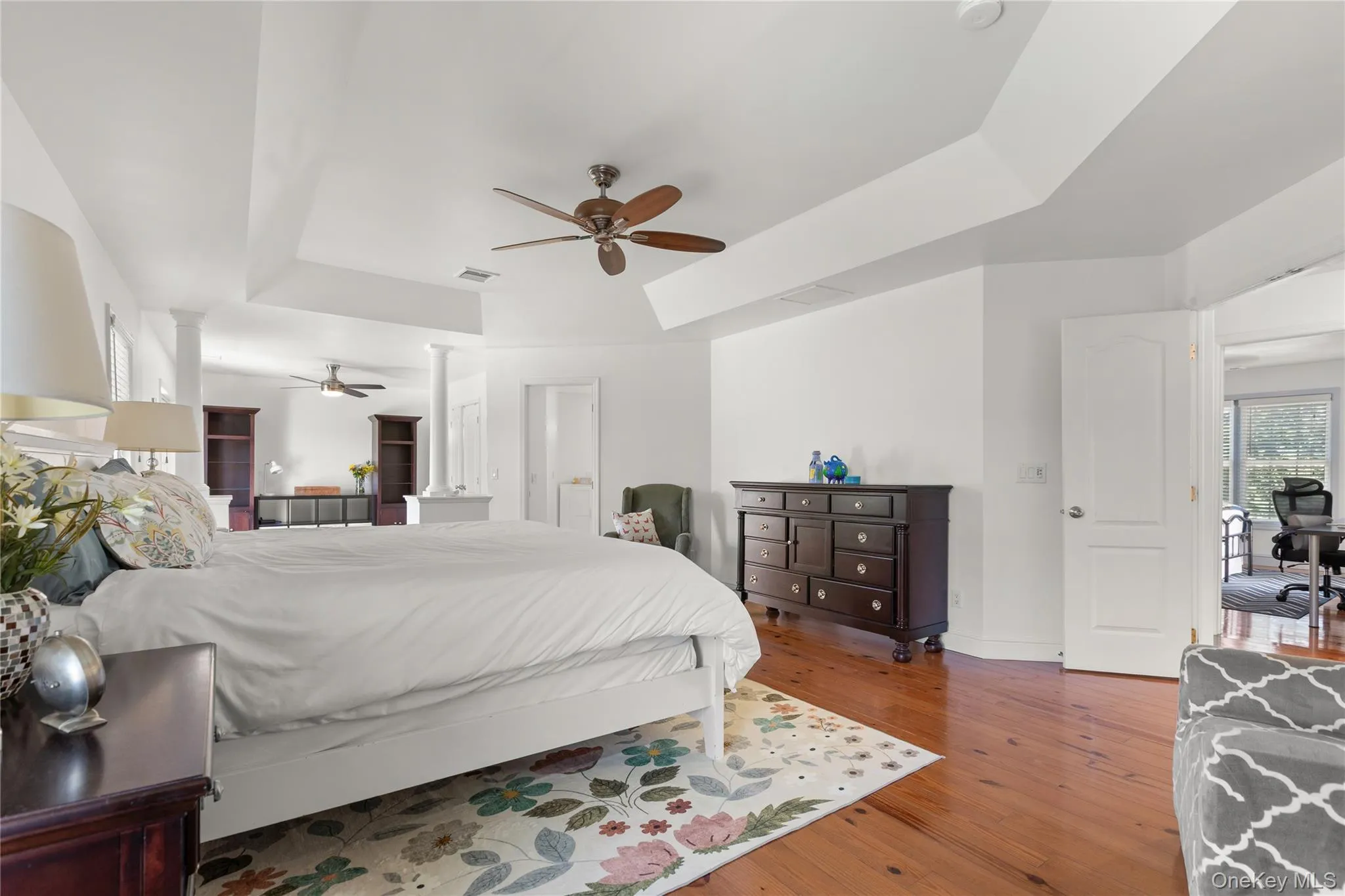 Spacious primary bedroom showcasing coffered ceiling architecture, sitting room with door to balcony, ceiling fan with wood blades, hardwood flooring throughout, and abundant natural light from large windows with plantation shutte Spacious primary bedroom showcasing coffered ceiling architecture, sitting room with door to balcony, ceiling fan with wood blades, hardwood flooring throughout, and abundant natural light from large windows with plantation shutte