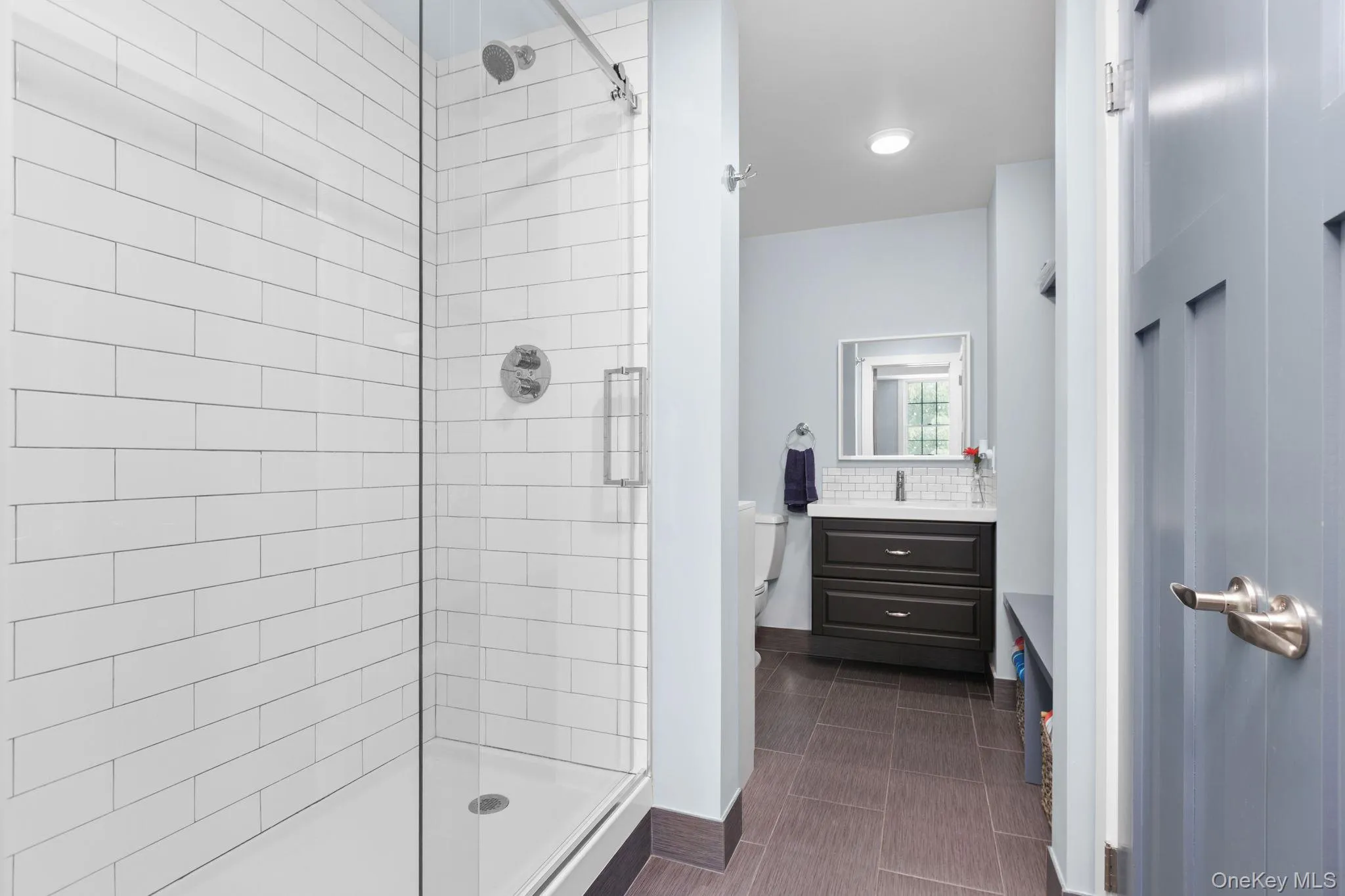 Bathroom featuring a walk-in shower with subway tile and frameless glass enclosure, single vanity with dark cabinetry recessed lighting, and tile flooring throughout Bathroom featuring a walk-in shower with subway tile and frameless glass enclosure, single vanity with dark cabinetry recessed lighting, and tile flooring throughout