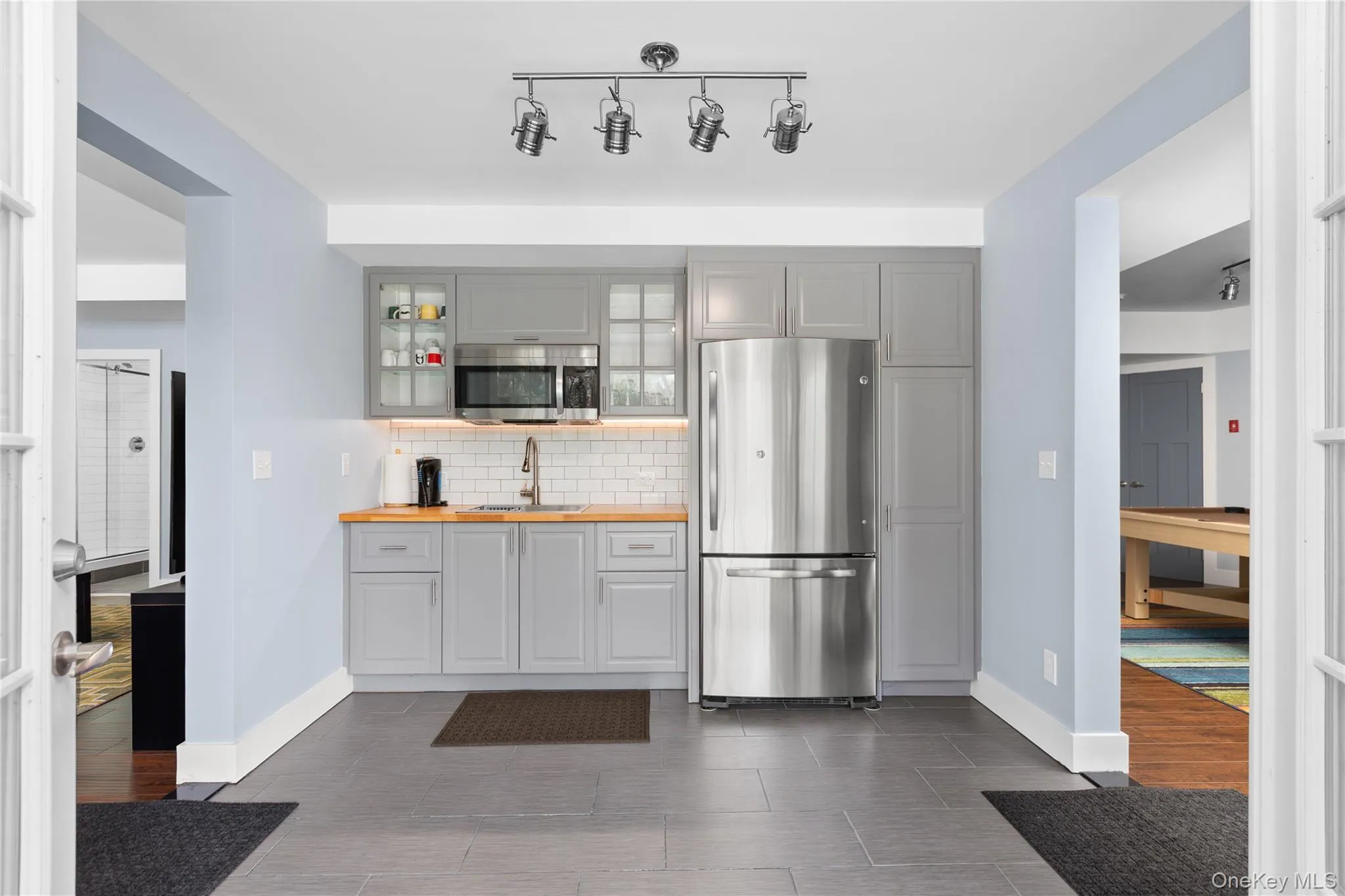 Kitchenette featuring gray cabinetry, stainless steel refrigerator and microwave, wood-finish countertops, subway tile backsplash, and track lighting. Kitchenette featuring gray cabinetry, stainless steel refrigerator and microwave, wood-finish countertops, subway tile backsplash, and track lighting.